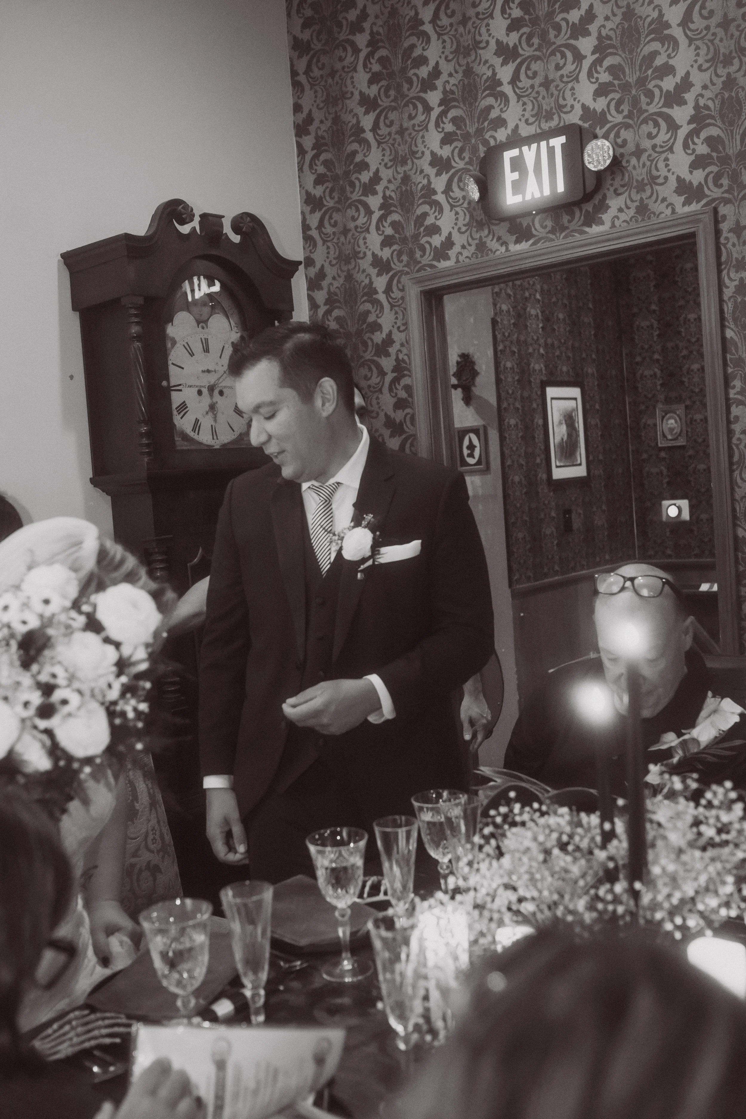 Groom standing and smiling during wedding reception while guests sit at candlelit table with floral arrangements