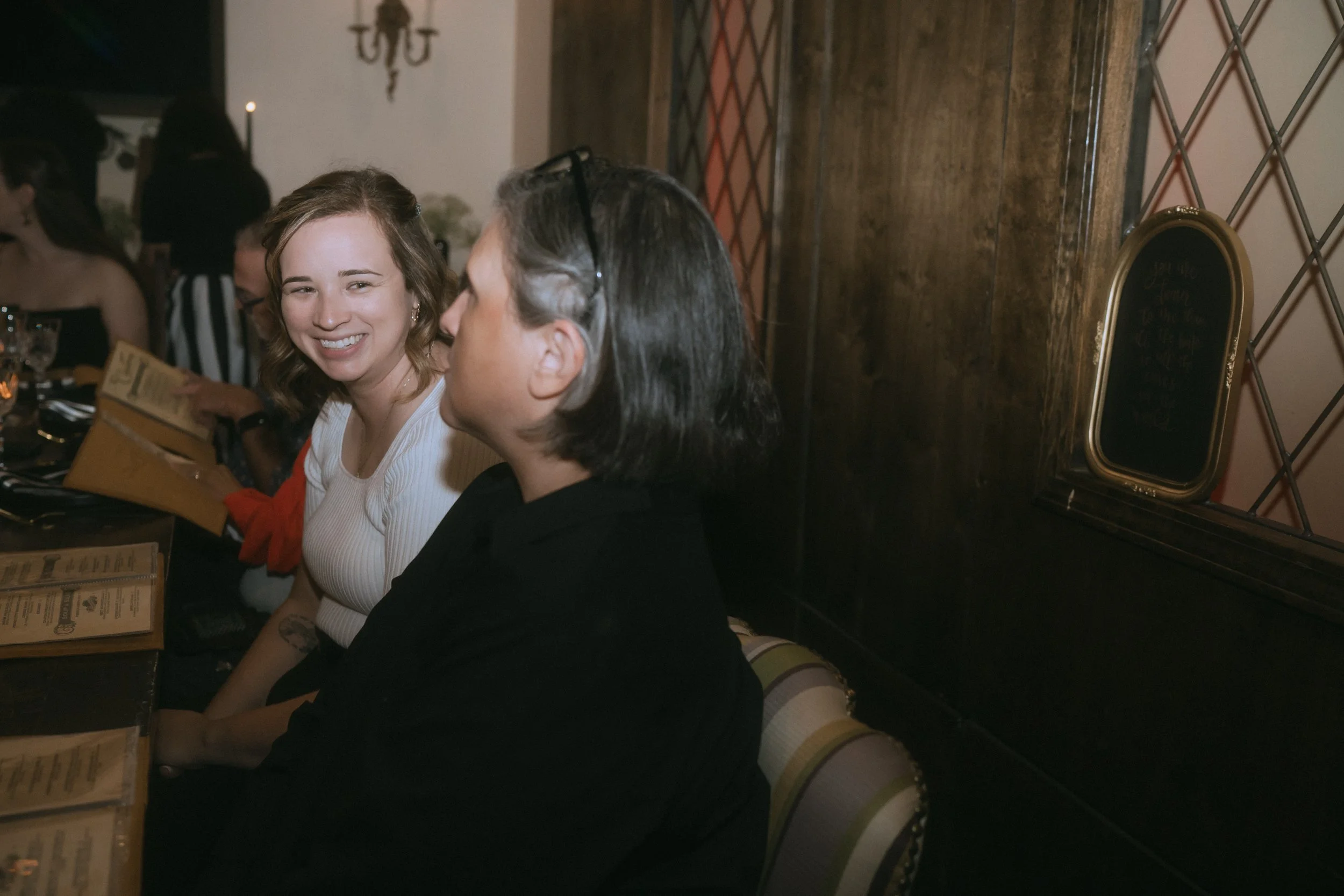 Wedding guests seated at reception table smiling and talking while looking at menus in dimly lit venue