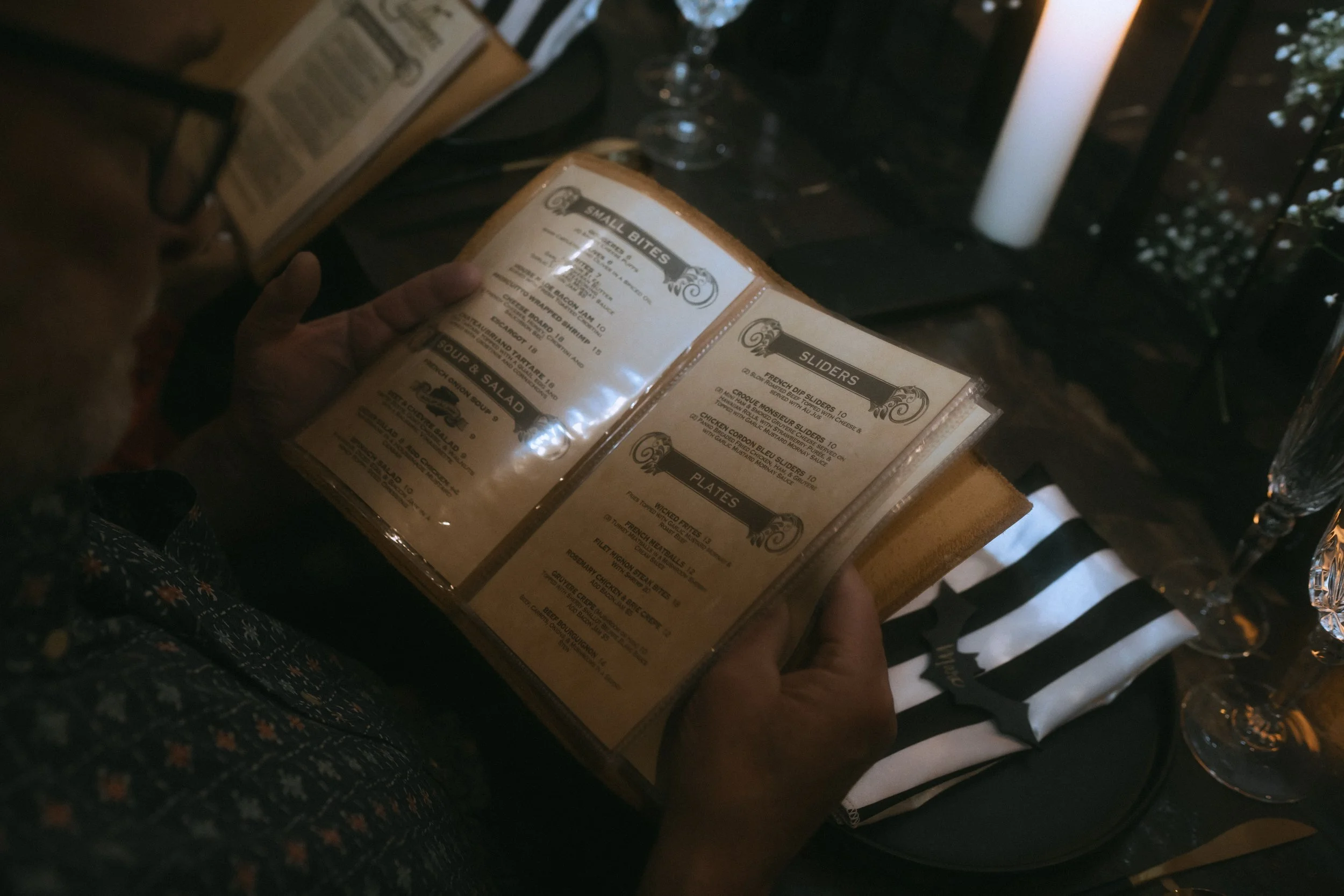 Close-up of guest holding wedding reception menu with candlelight, glassware, and striped napkin on table