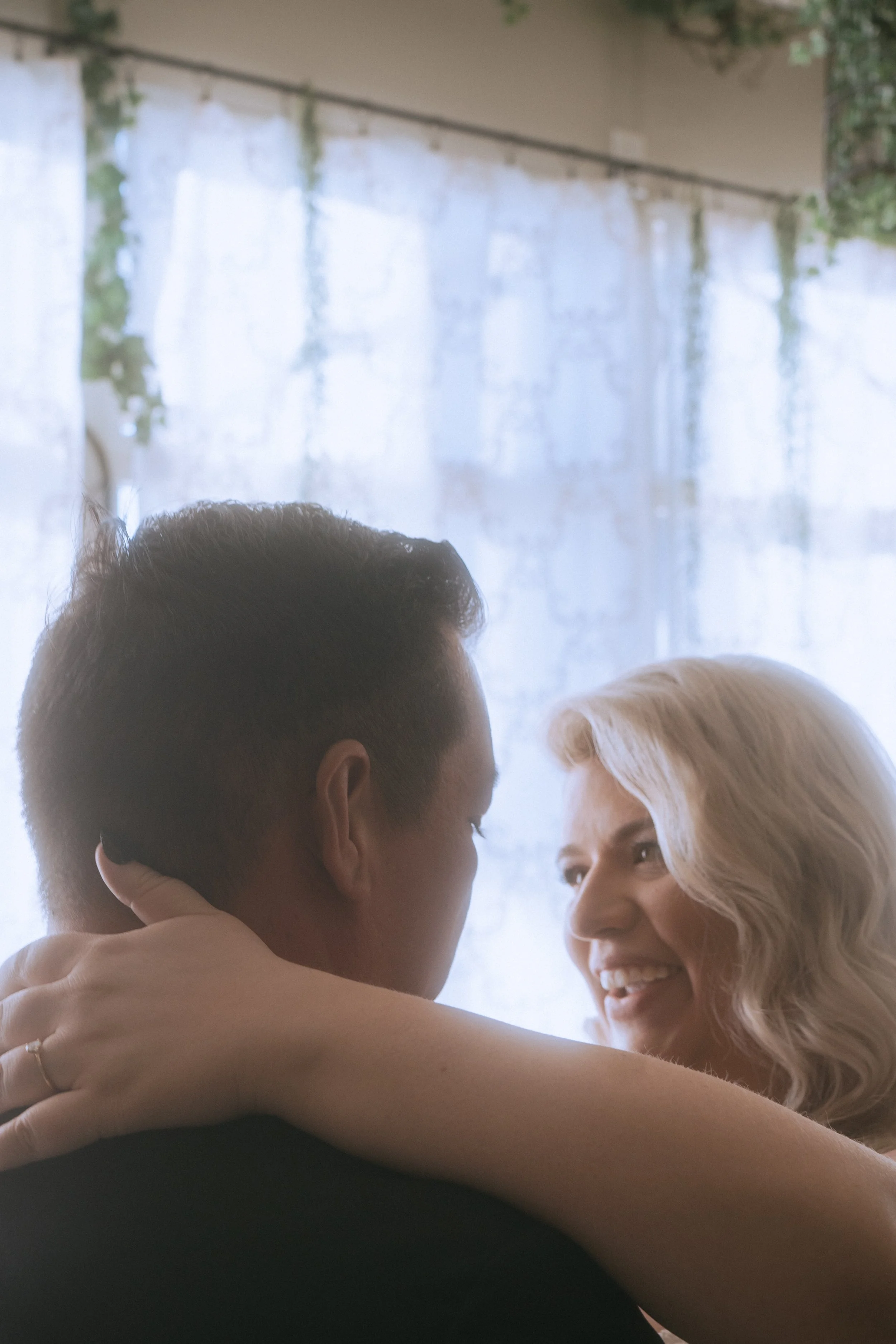 Bride smiling while embracing groom in soft natural window light during intimate wedding moment