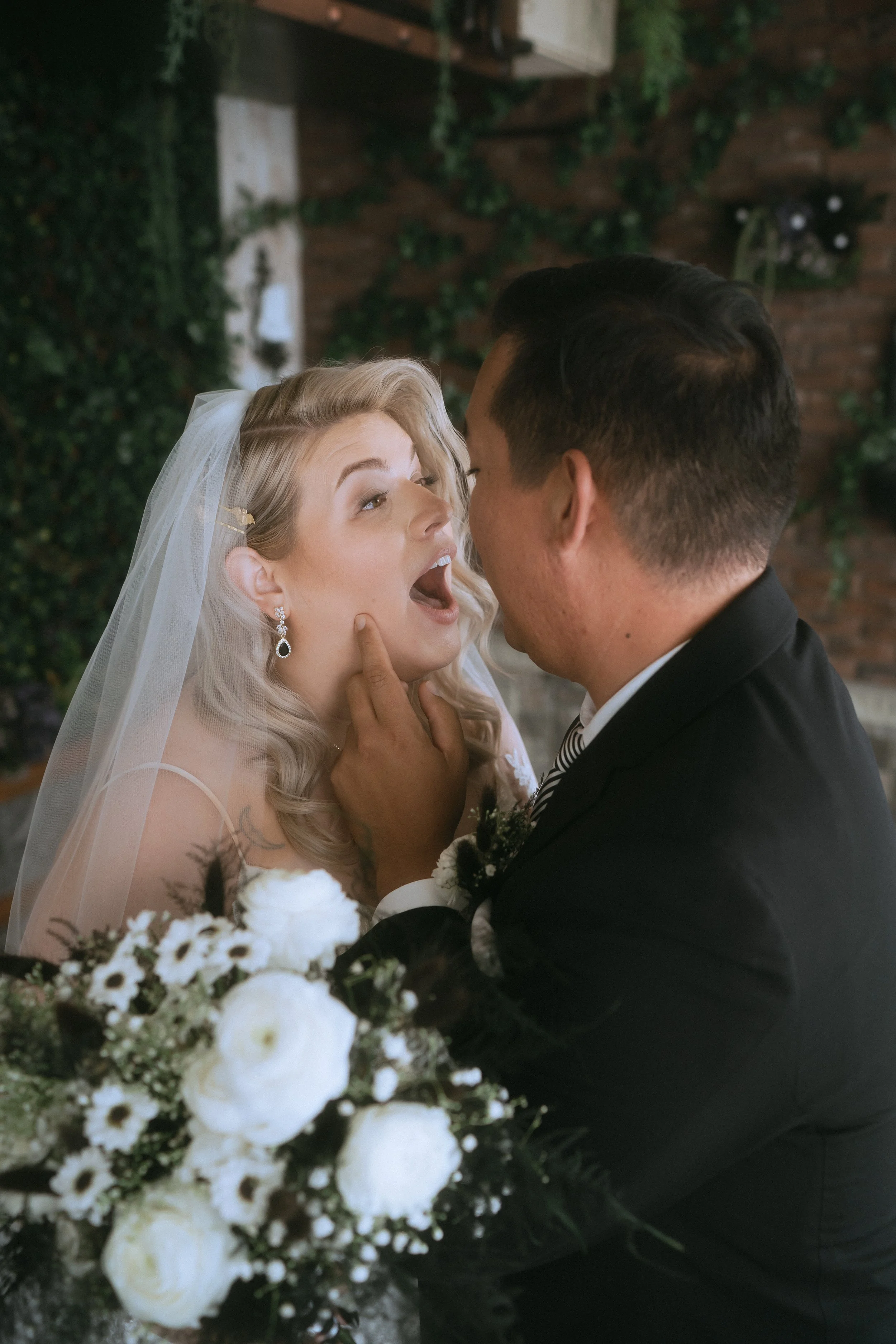 Bride laughing as groom playfully feeds cake during wedding reception with greenery backdrop