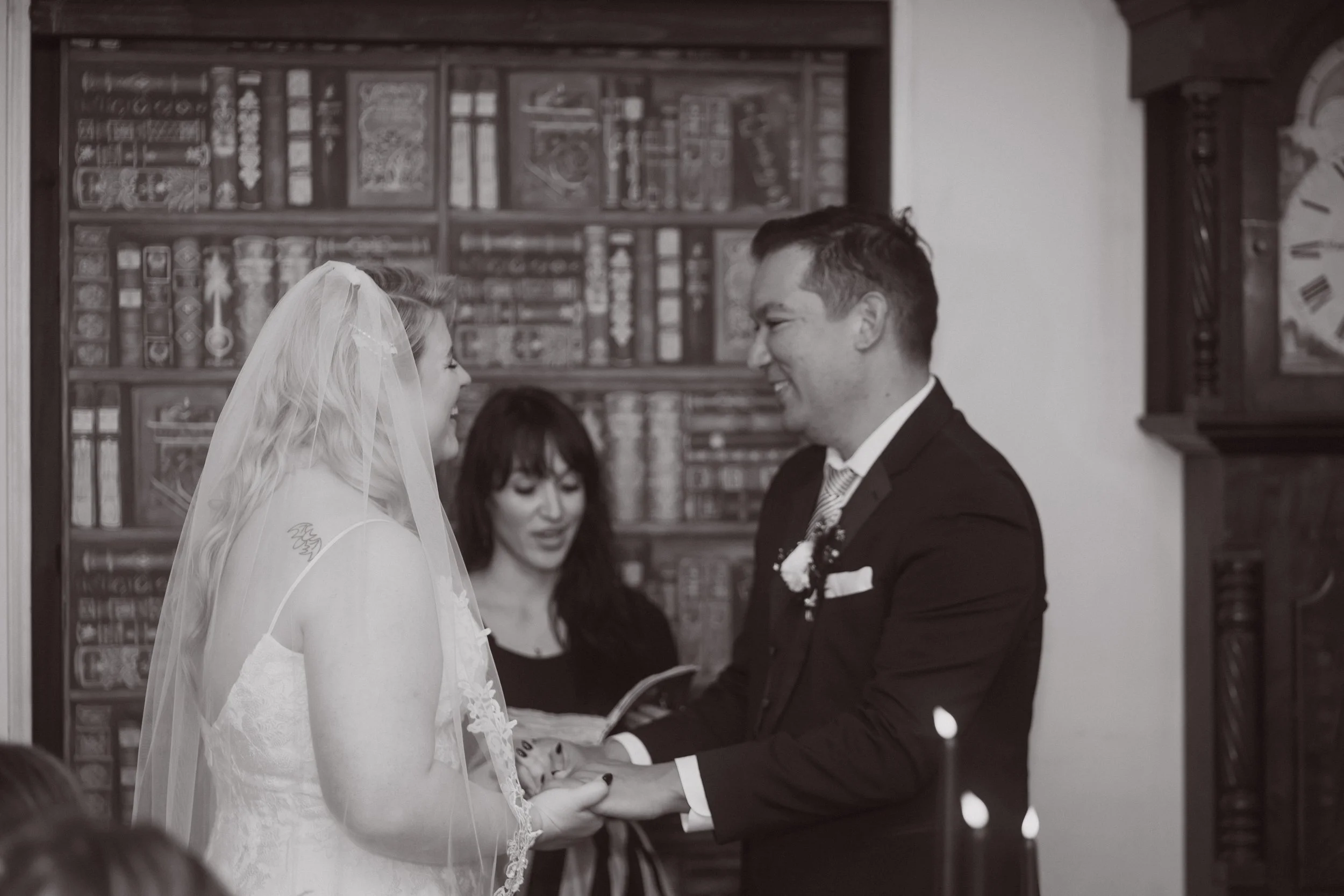 Bride and groom holding hands and smiling during wedding ceremony while exchanging vows with officiant in background