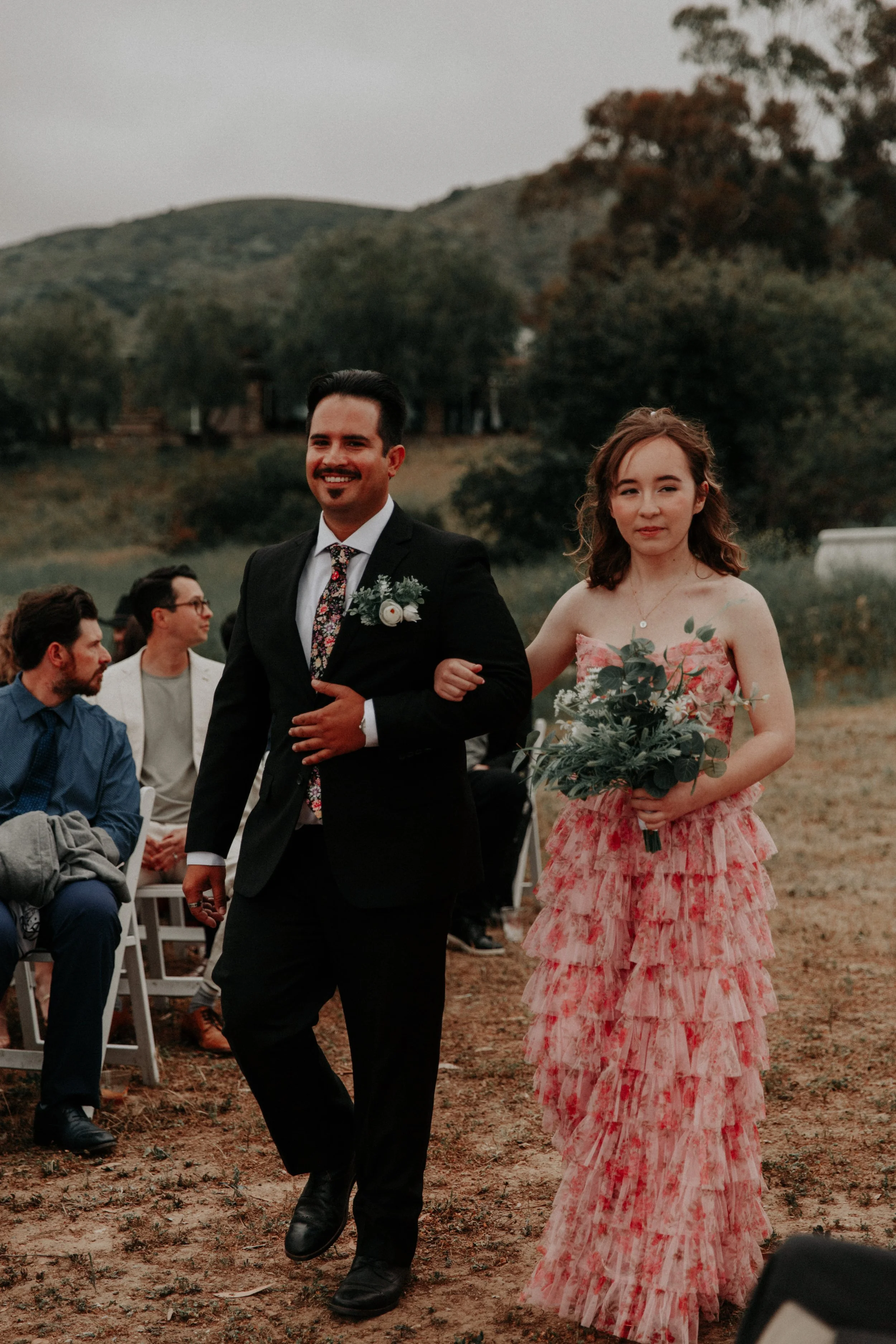 Groomsman and bridesmaid walking together down outdoor wedding ceremony aisle holding bouquet
