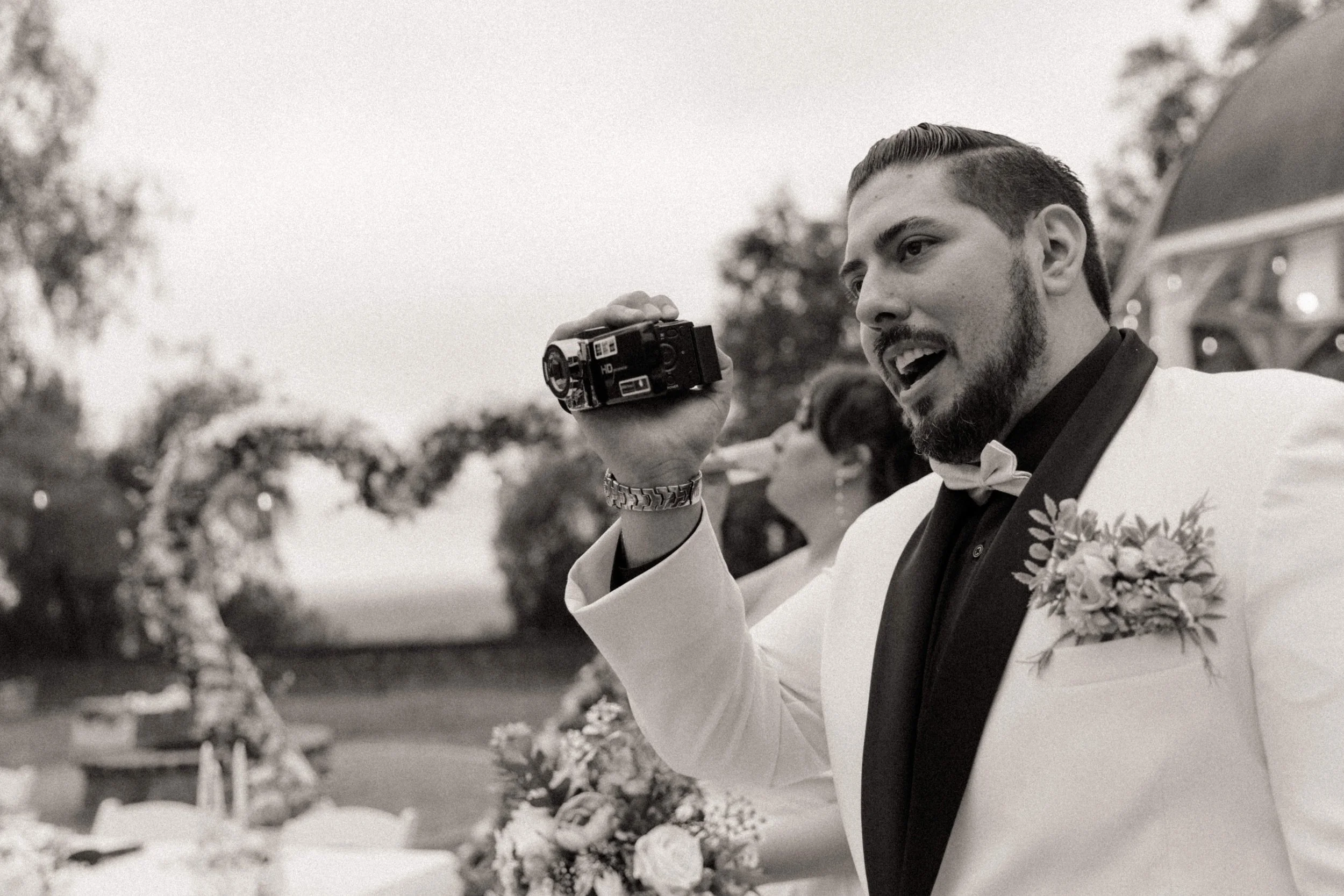 Groom holding vintage camcorder during playful alternative wedding reception with floral heart arch in background