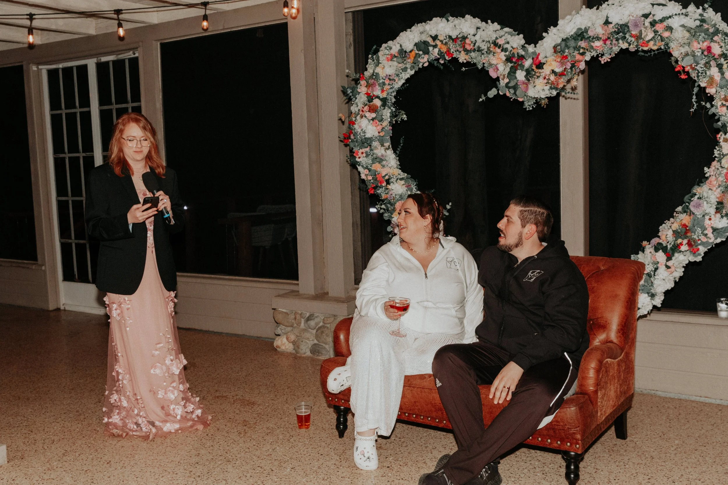 Wedding speech moment with newlyweds seated beneath oversized floral heart installation during reception