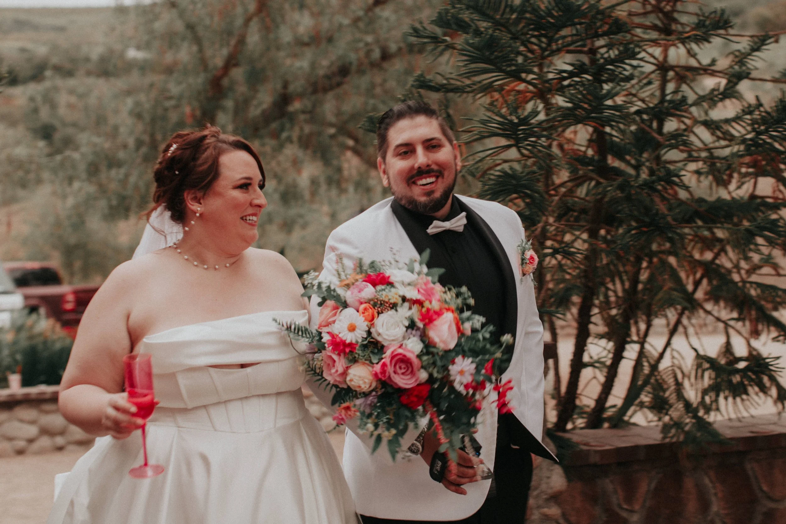 Smiling bride and groom entering wedding reception holding colorful bouquet and champagne glass