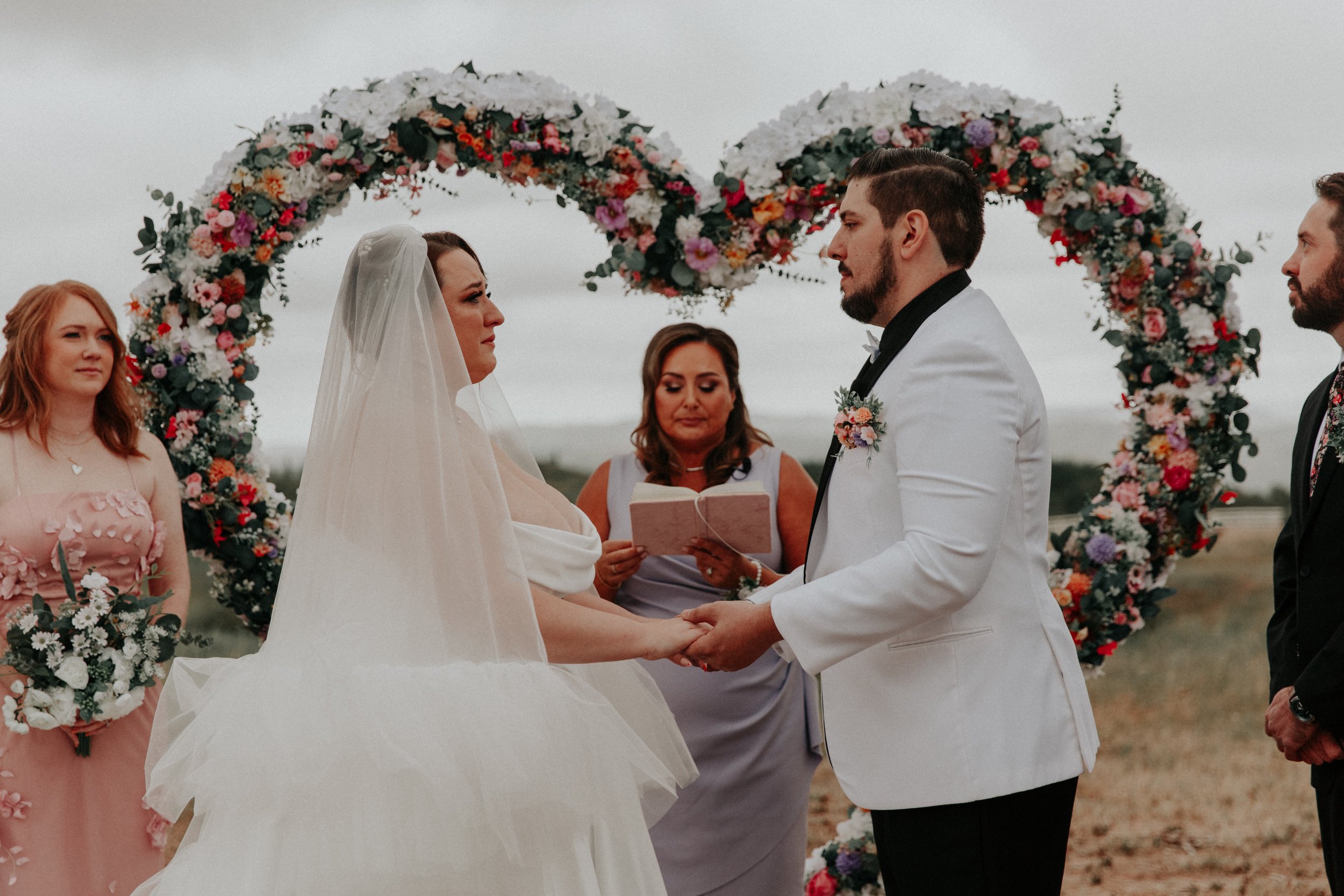 Bride and groom holding hands beneath floral heart arches during wedding ceremony
