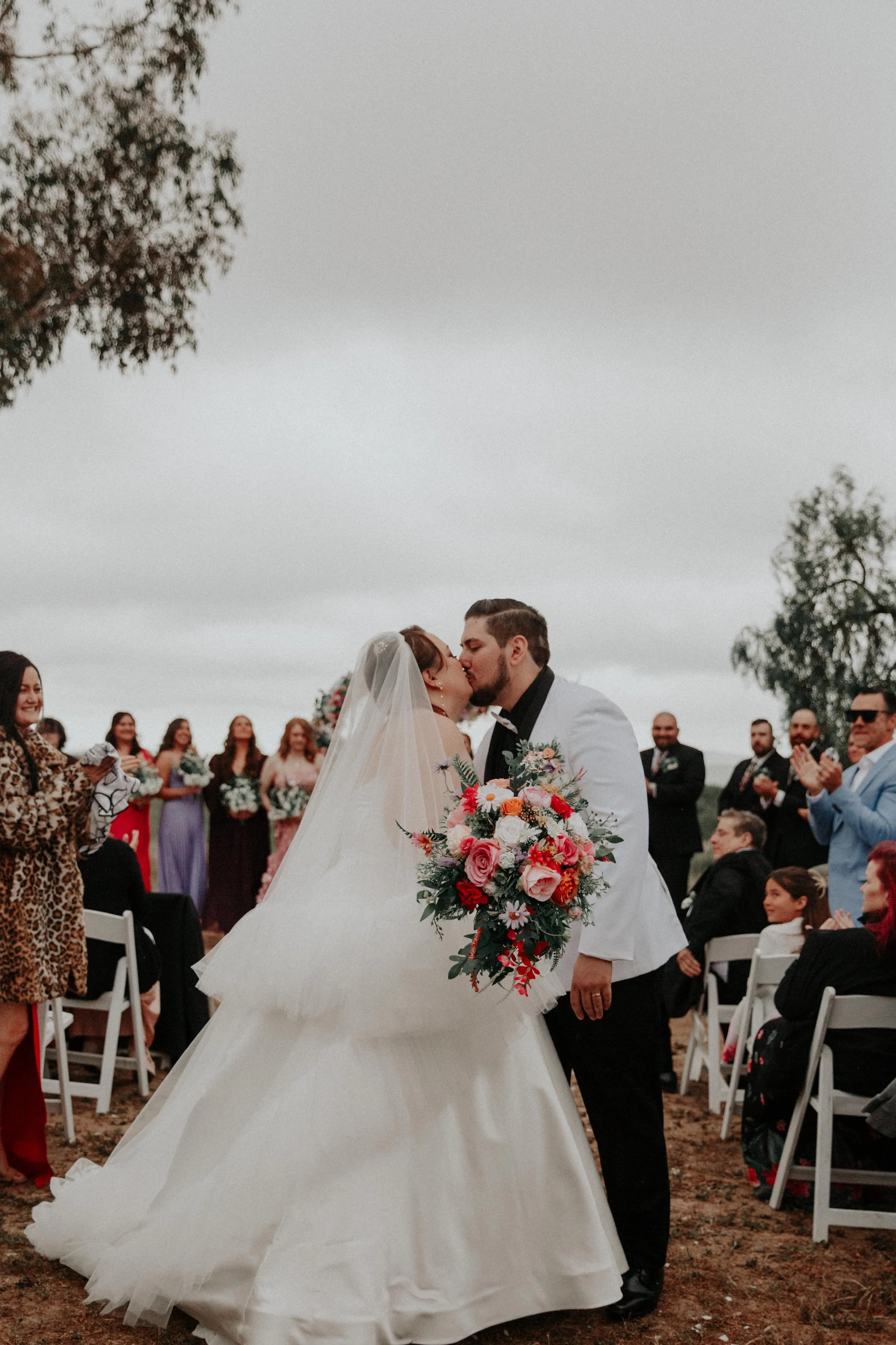 Bride and groom sharing first kiss outdoors with mountain landscape and wedding party nearby
