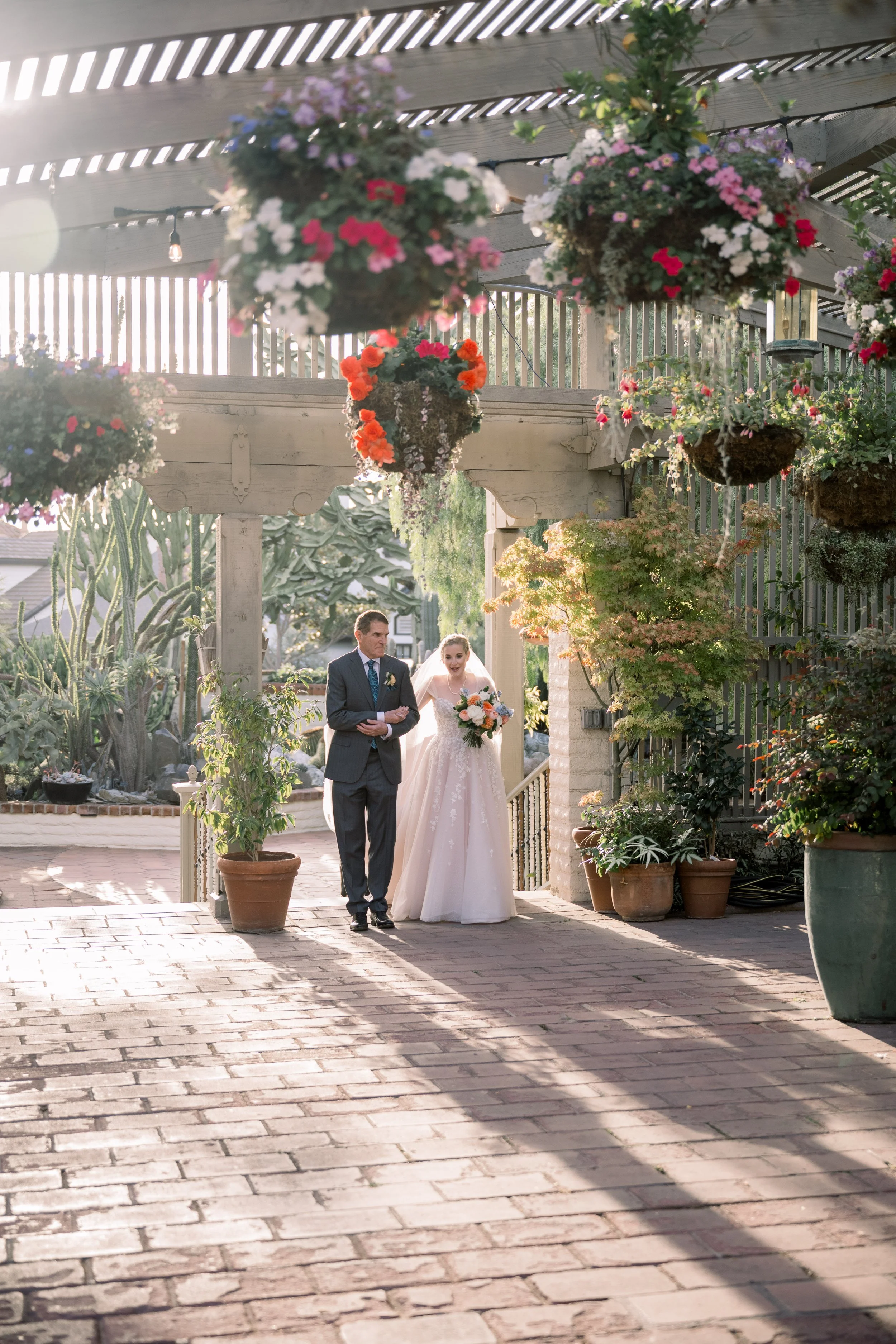 Bride walking arm in arm with her father toward the ceremony space beneath hanging floral baskets and warm sunlight