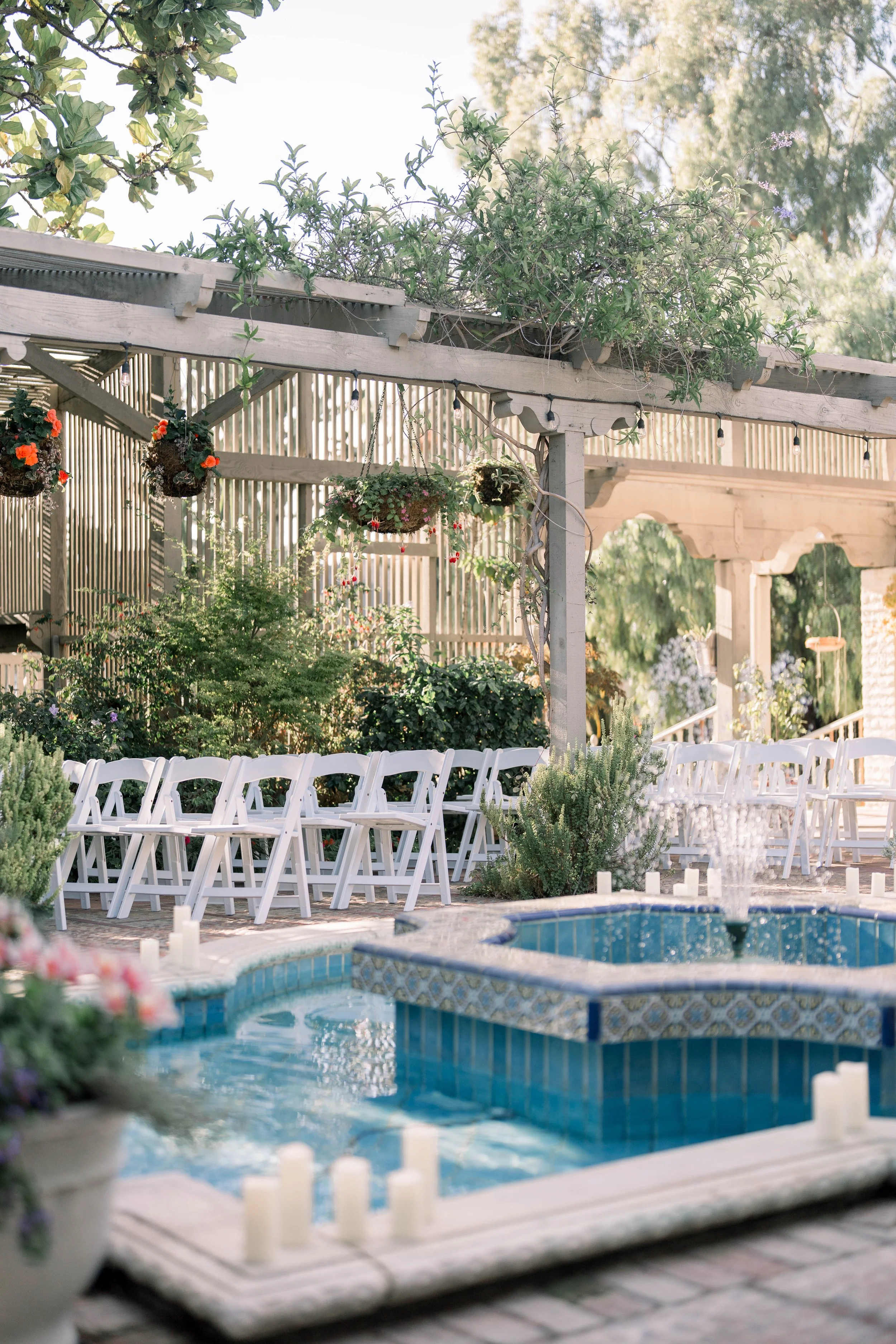 White ceremony chairs arranged beside a tiled fountain with hanging floral baskets and lush greenery overhead