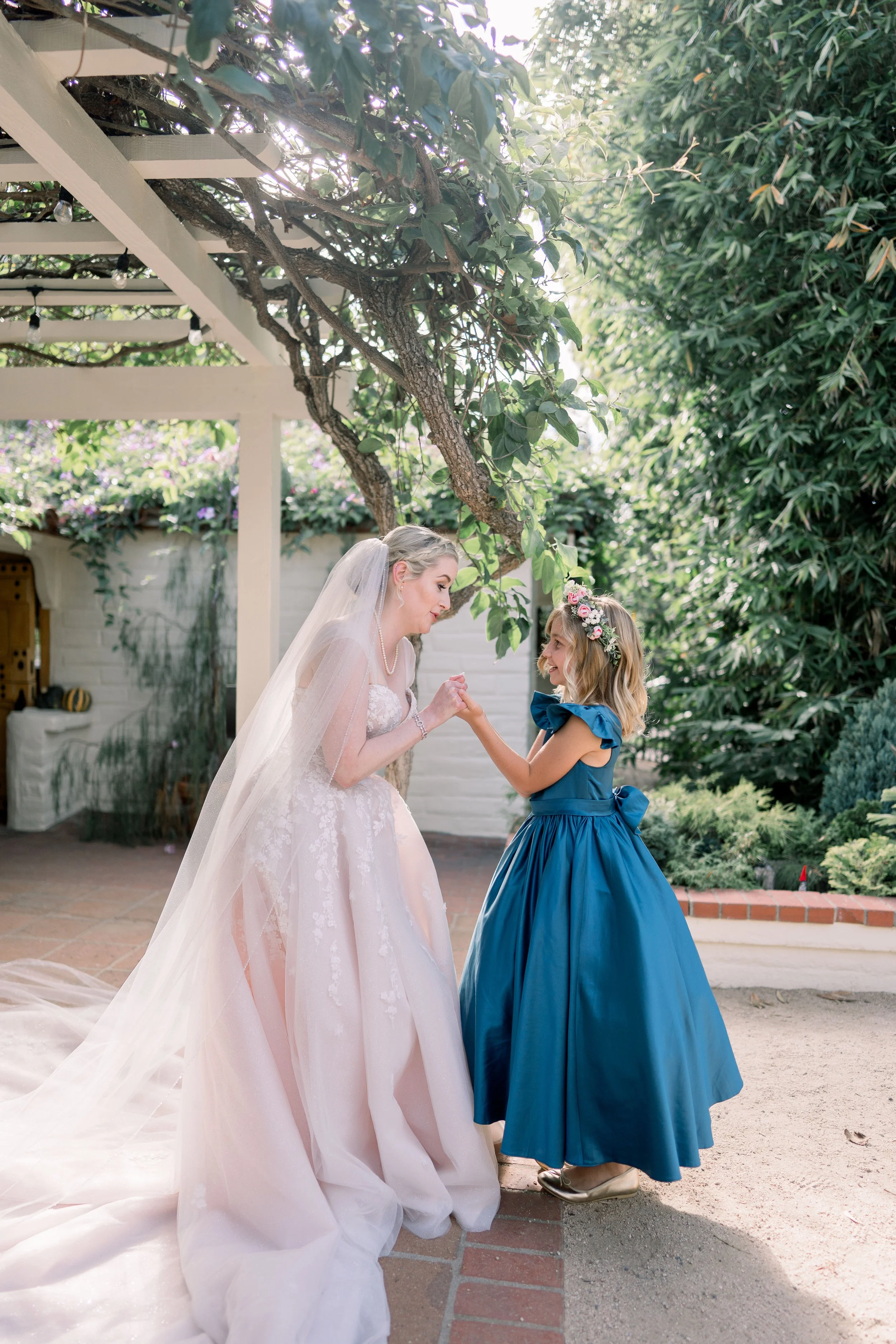 Bride holding hands with a flower girl in a blue dress beneath a garden pergola during wedding portraits