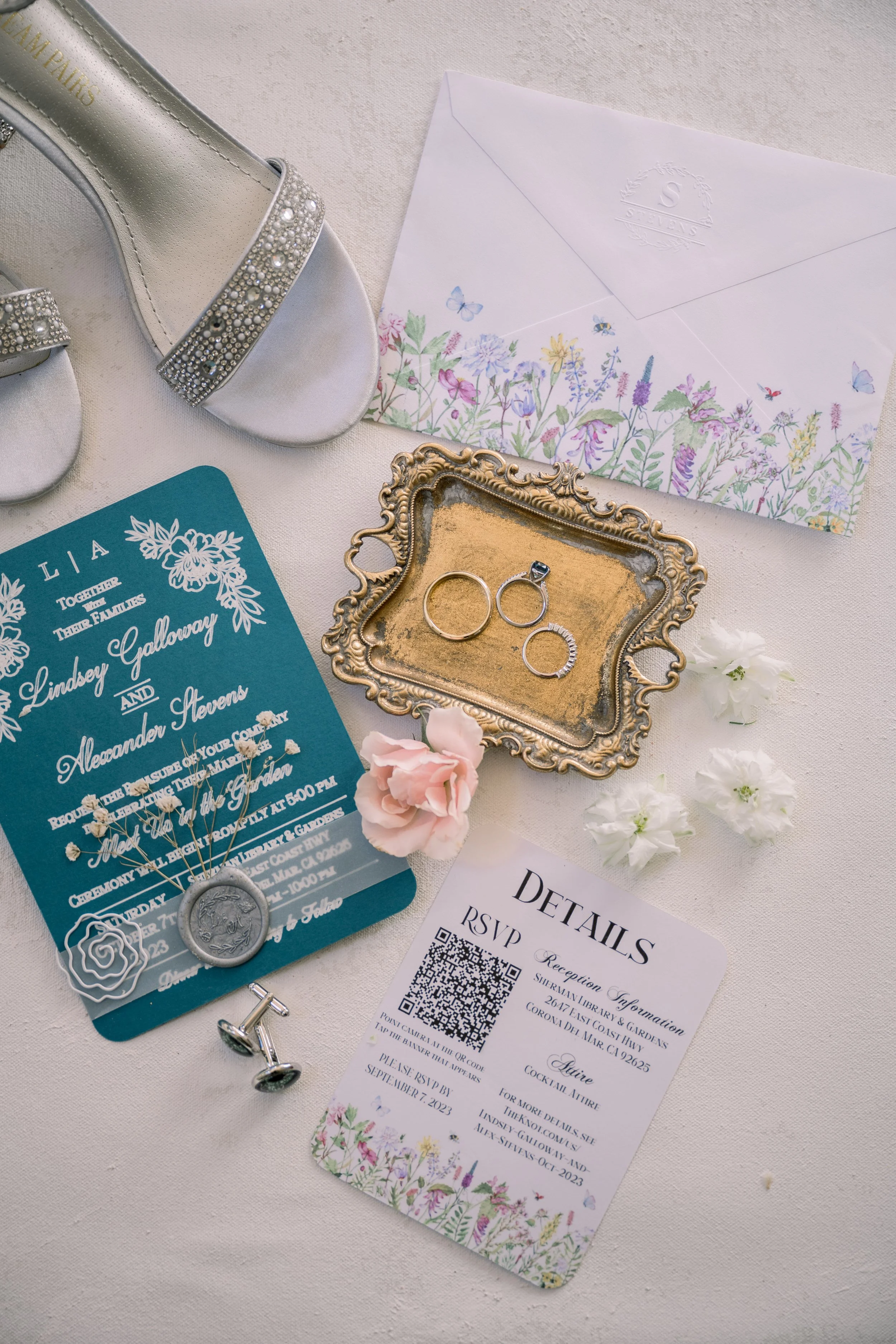 Wedding flat lay featuring invitations, rings, cufflinks, floral details, and bridal shoes arranged on fabric