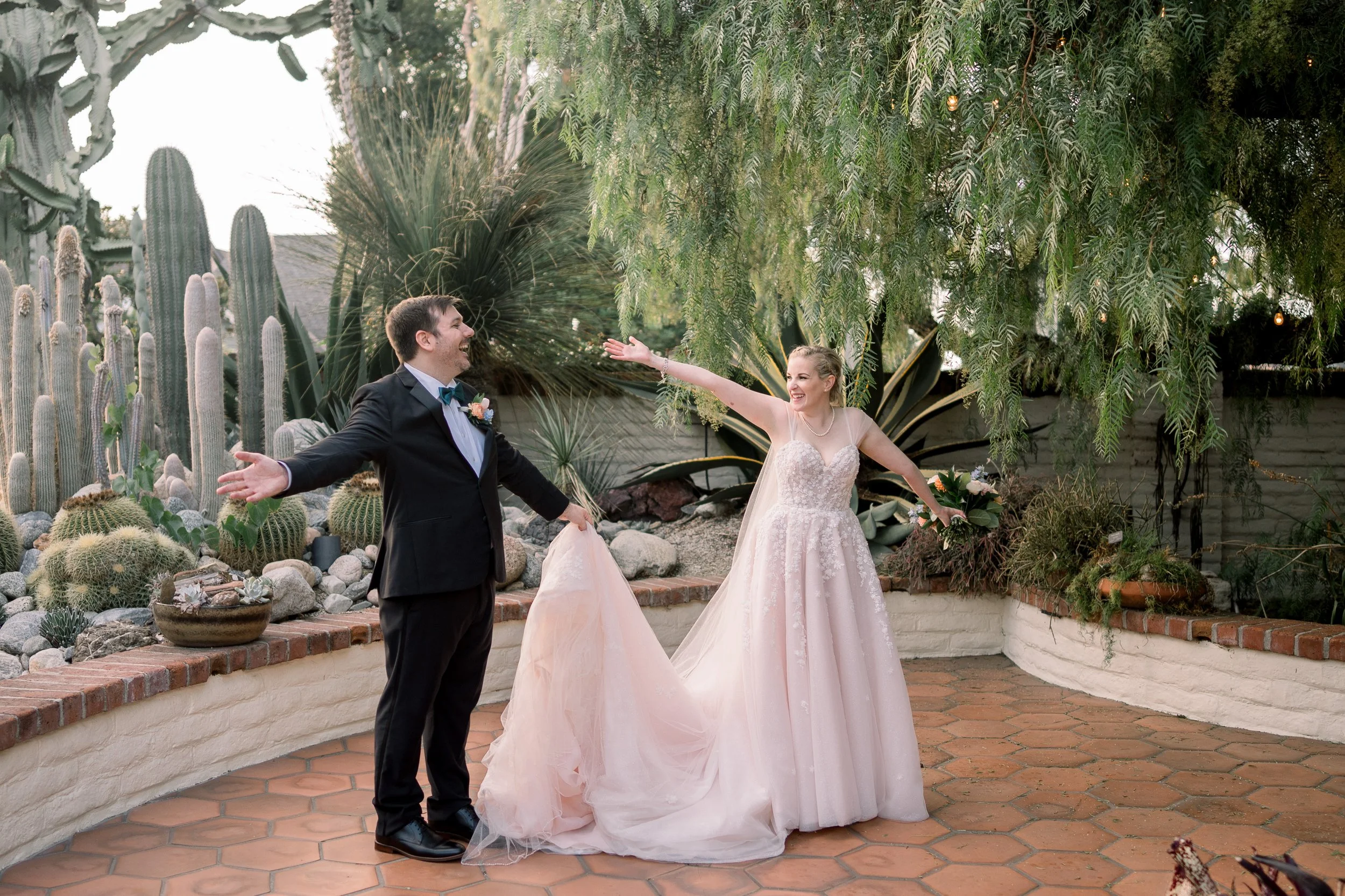 Bride and groom posing playfully with outstretched arms during wedding portraits in a garden filled with cacti and greenery