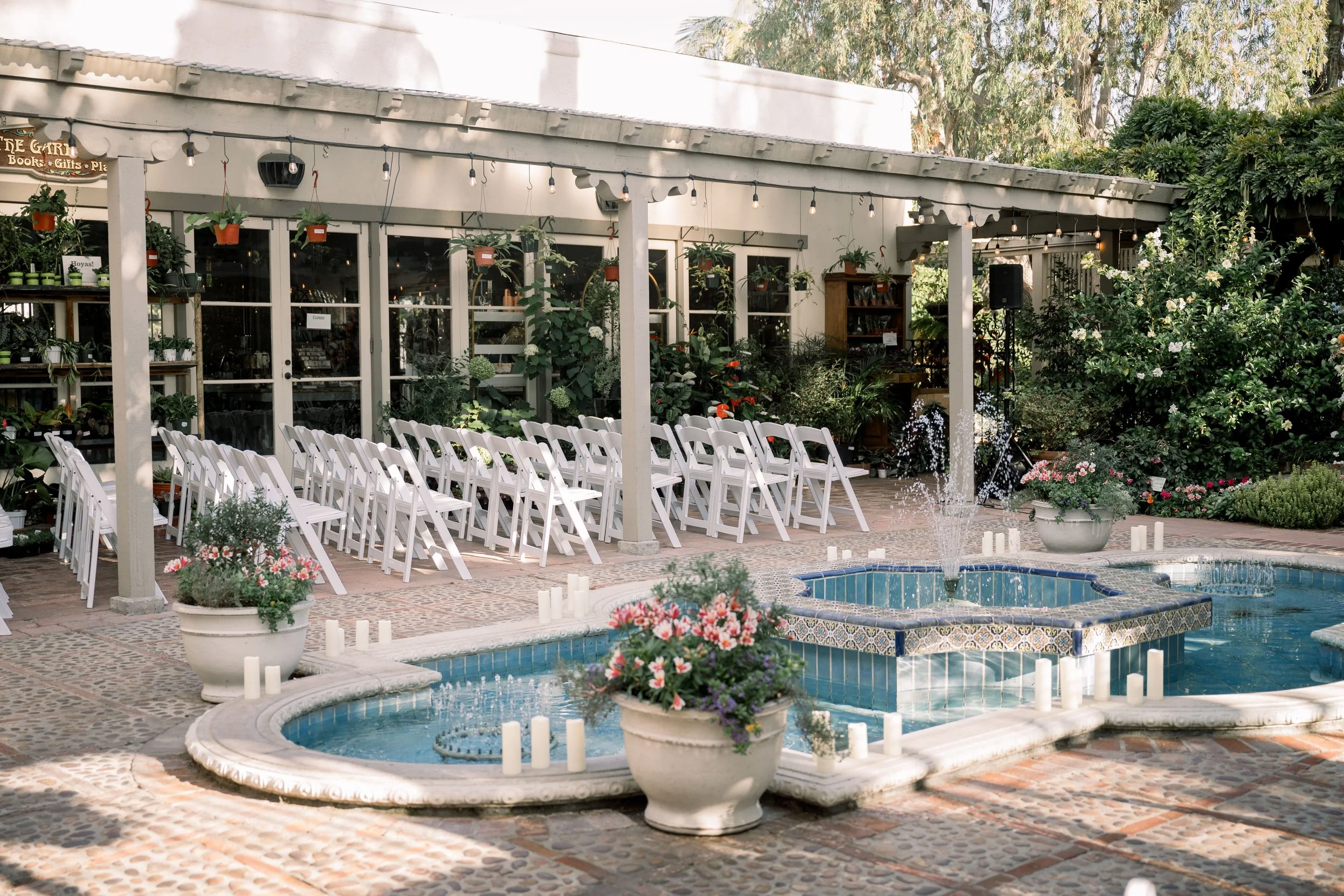 Outdoor wedding ceremony setup with white folding chairs surrounding a tiled fountain courtyard and garden-inspired decor