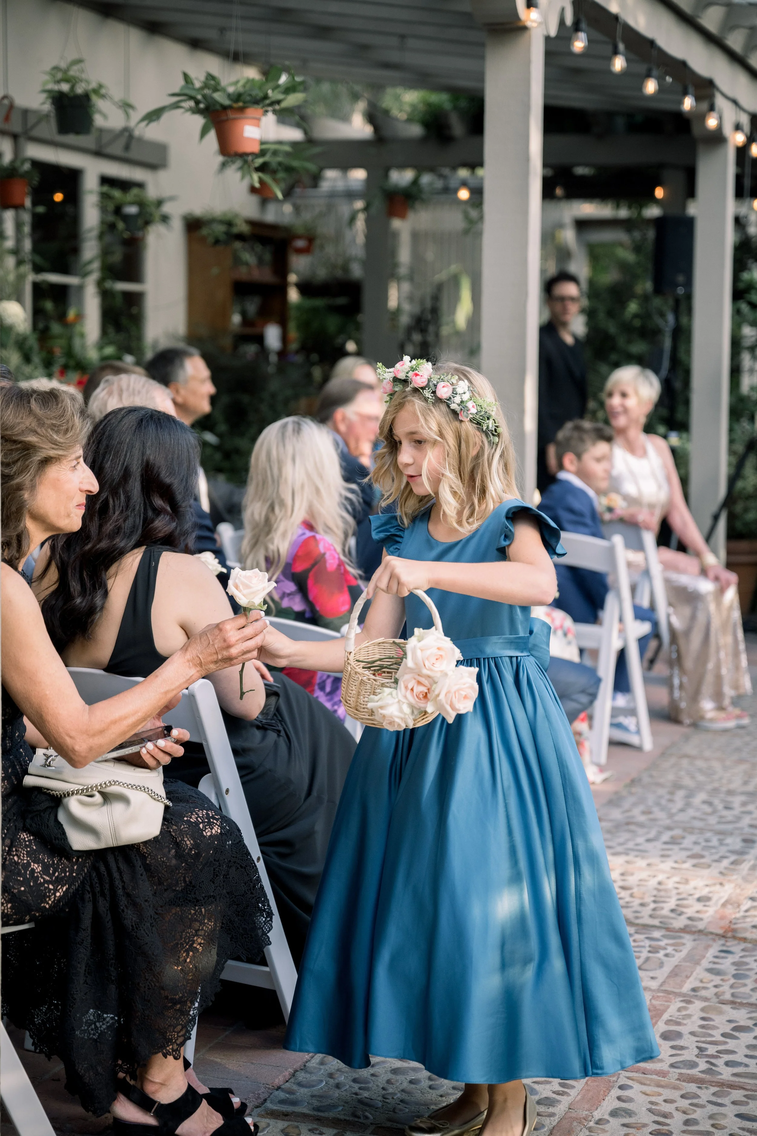 Flower girl in a blue dress and floral crown handing out roses to wedding guests during the ceremony