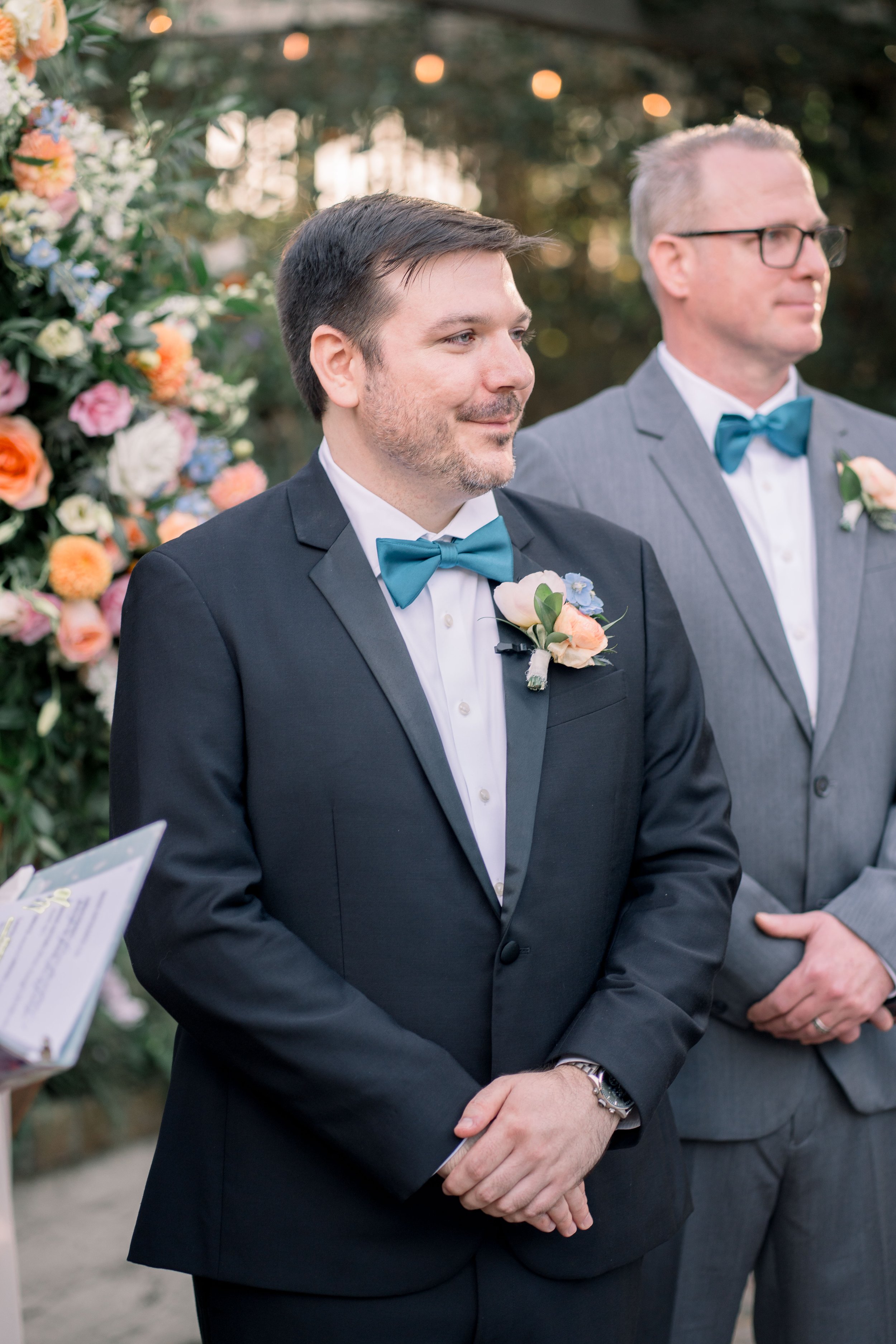 Groom smiling during the wedding ceremony while standing beside his best man and floral altar decor