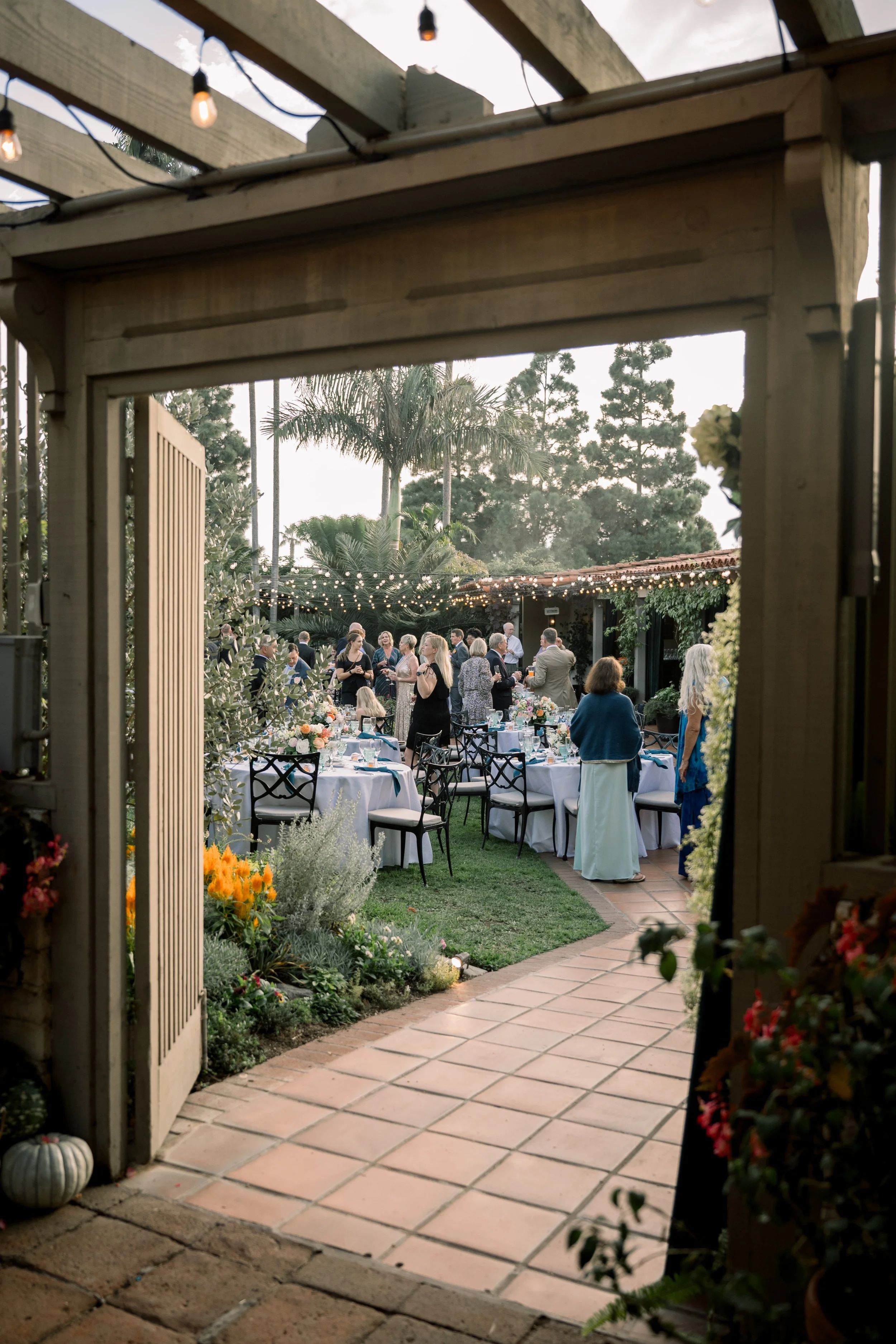 View into an outdoor garden wedding reception with guests mingling beneath string lights and floral tablescapes