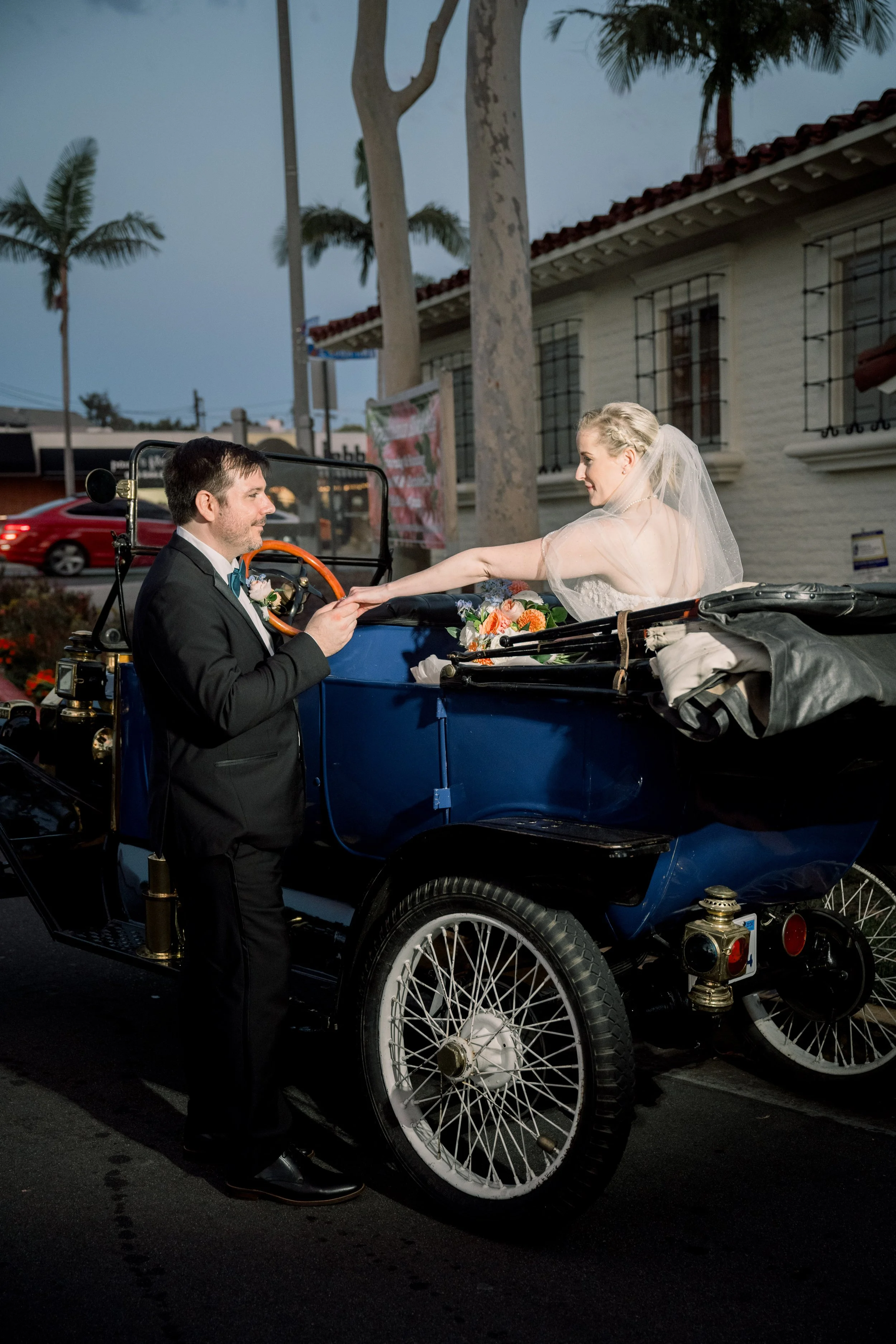 Bride reaching for the groom’s hand while sitting in a vintage convertible getaway car after the wedding celebration