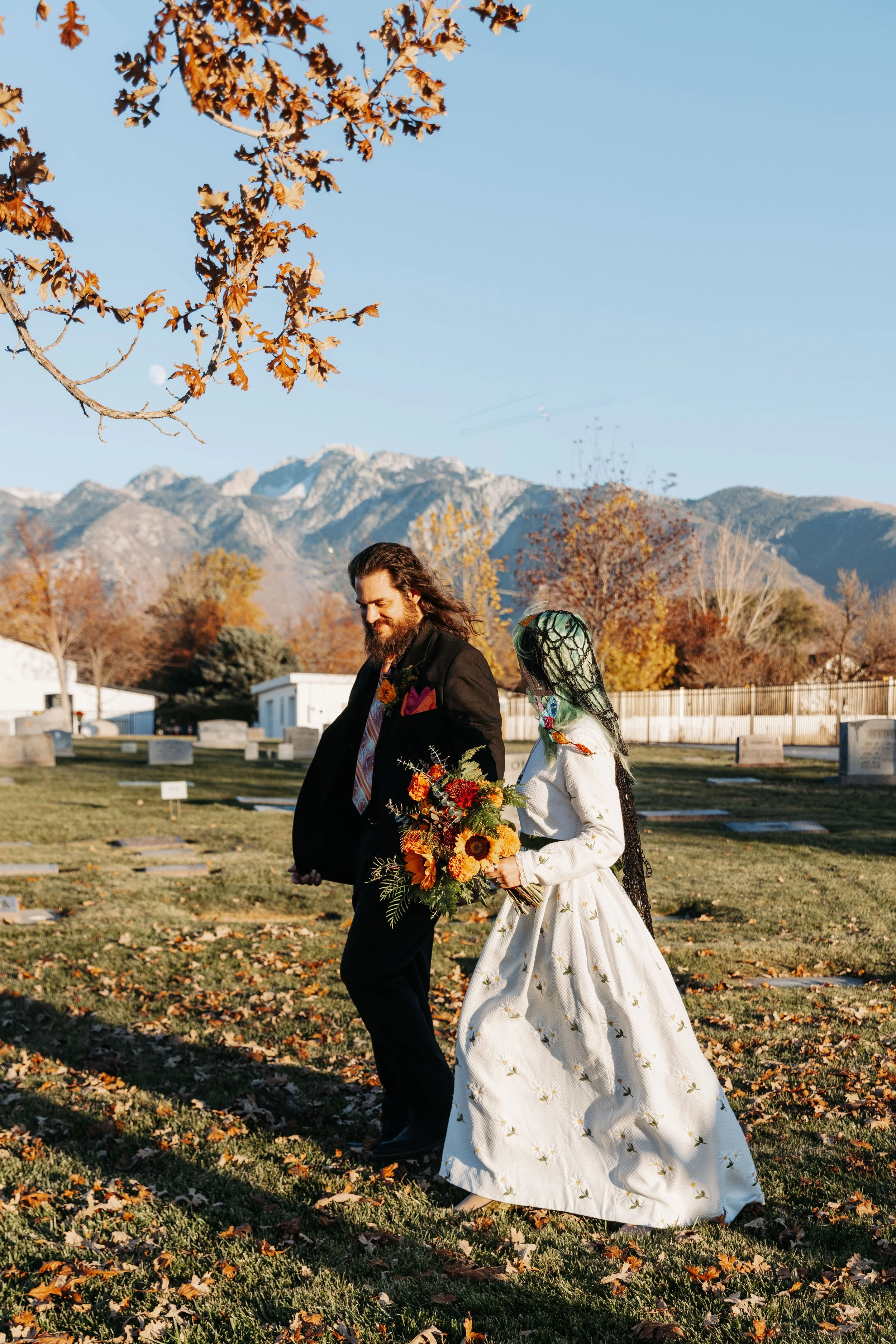 Bride and groom walk hand in hand through a cemetery with fall foliage and mountain views, creating meaningful portraits that reflected their unique wedding vision.