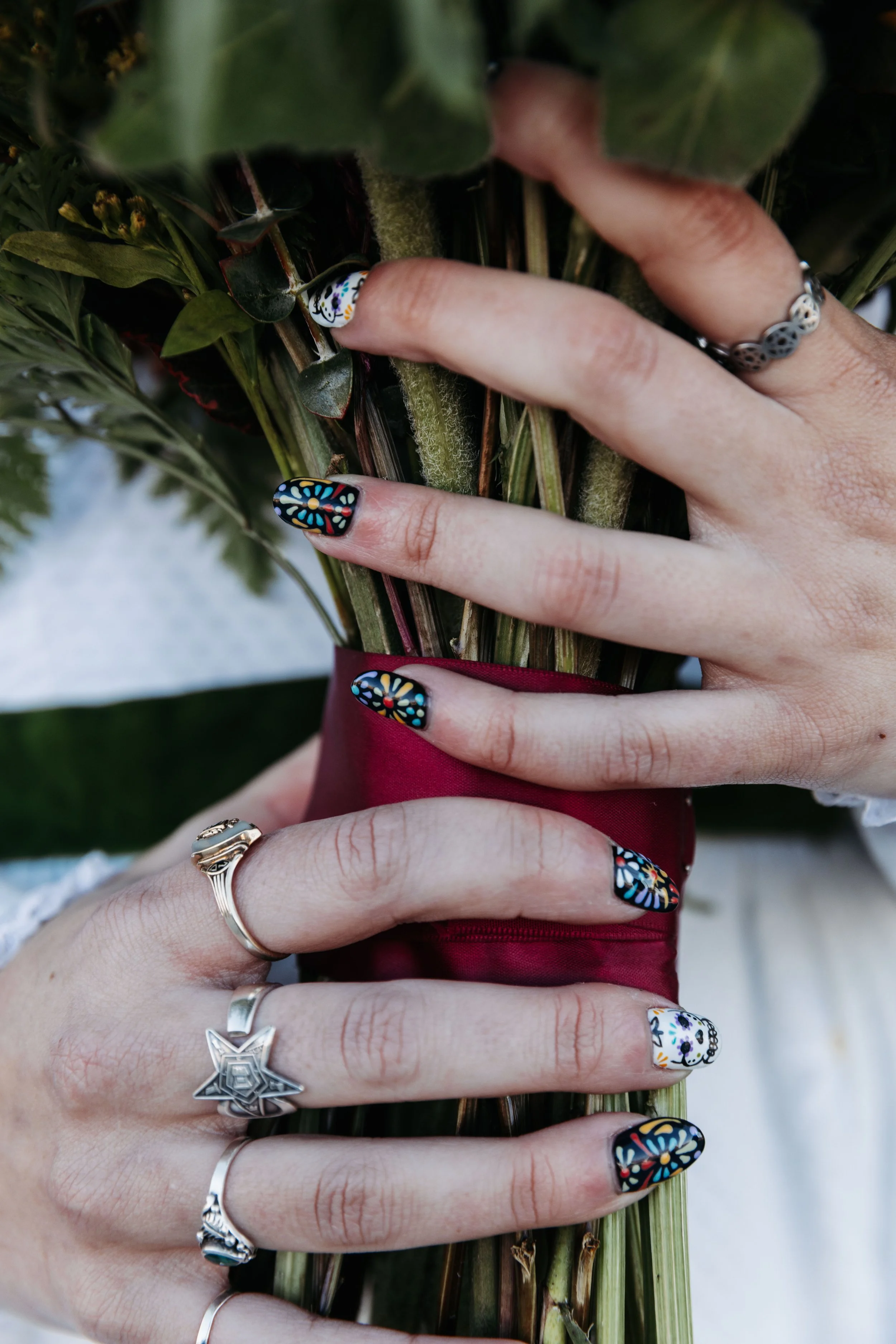 Close-up of the bride's hands holding her bouquet, showcasing colorful Day of the Dead-inspired nail art, silver rings, and burgundy ribbon wrapping.