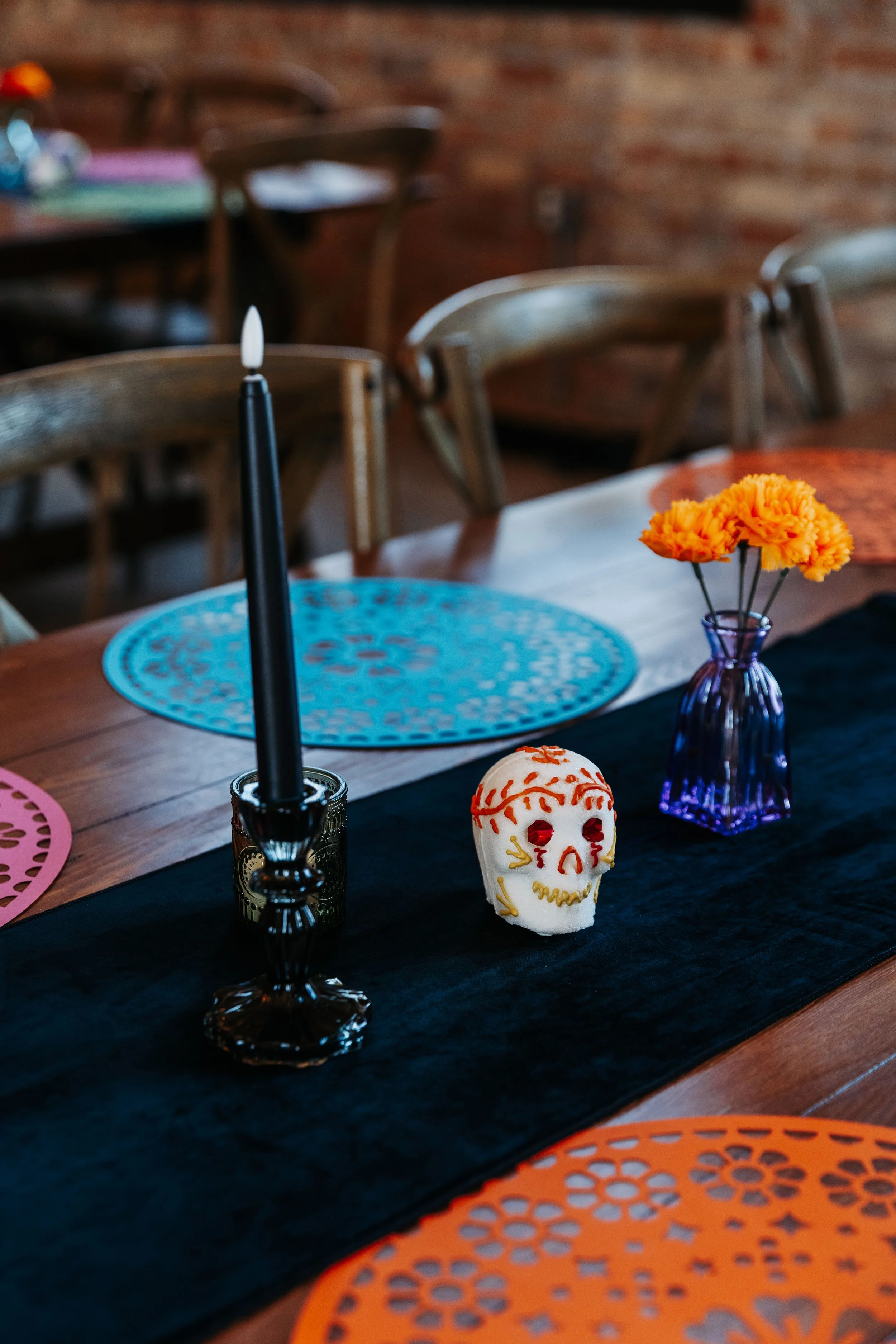 Reception table decorated with colorful papel picado placemats, marigolds, black taper candles, and sugar skull accents for a Día de los Muertos wedding theme.