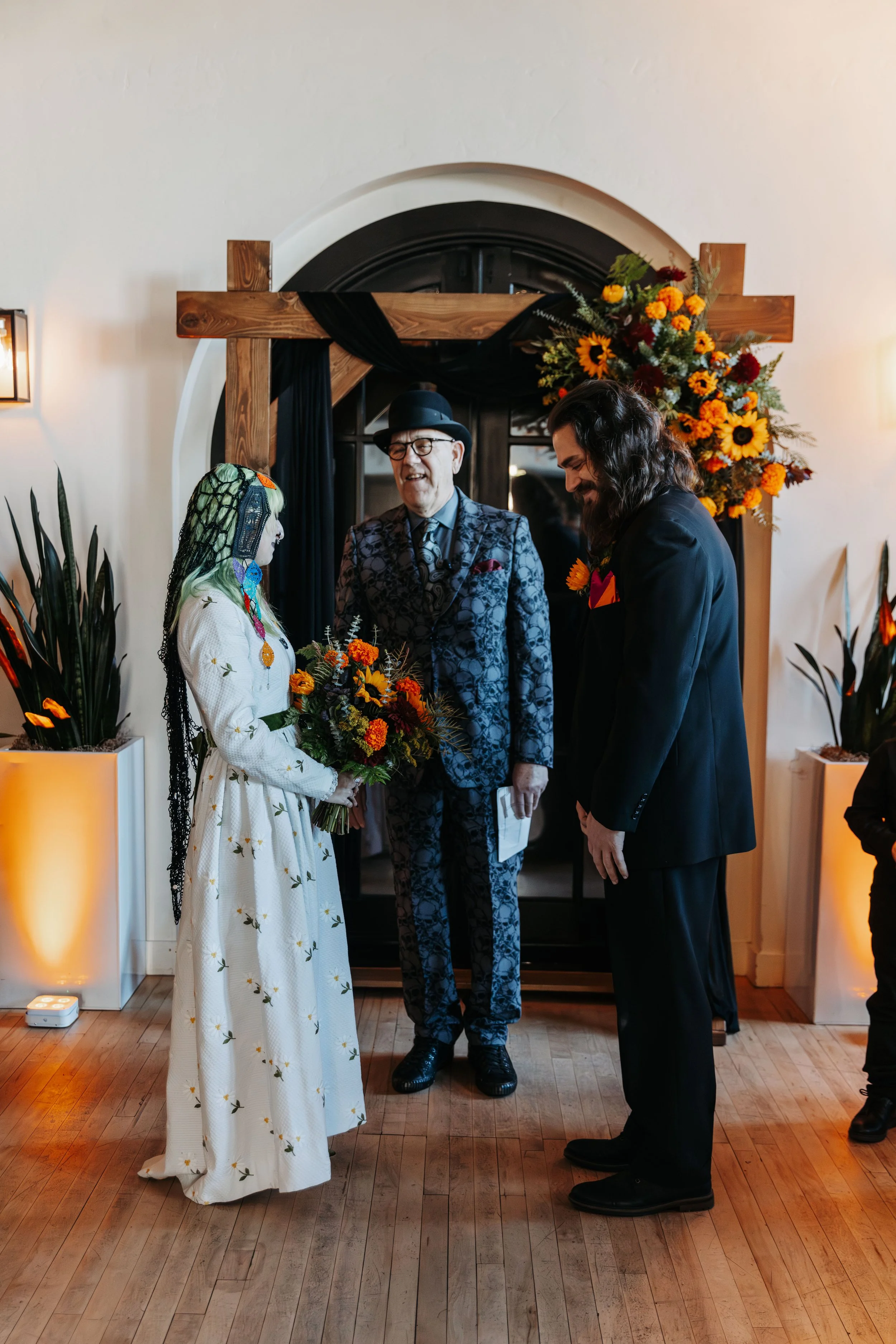 Bride and groom stand together during their wedding ceremony beneath a wooden altar adorned with black draping and vibrant sunflower floral arrangements.
