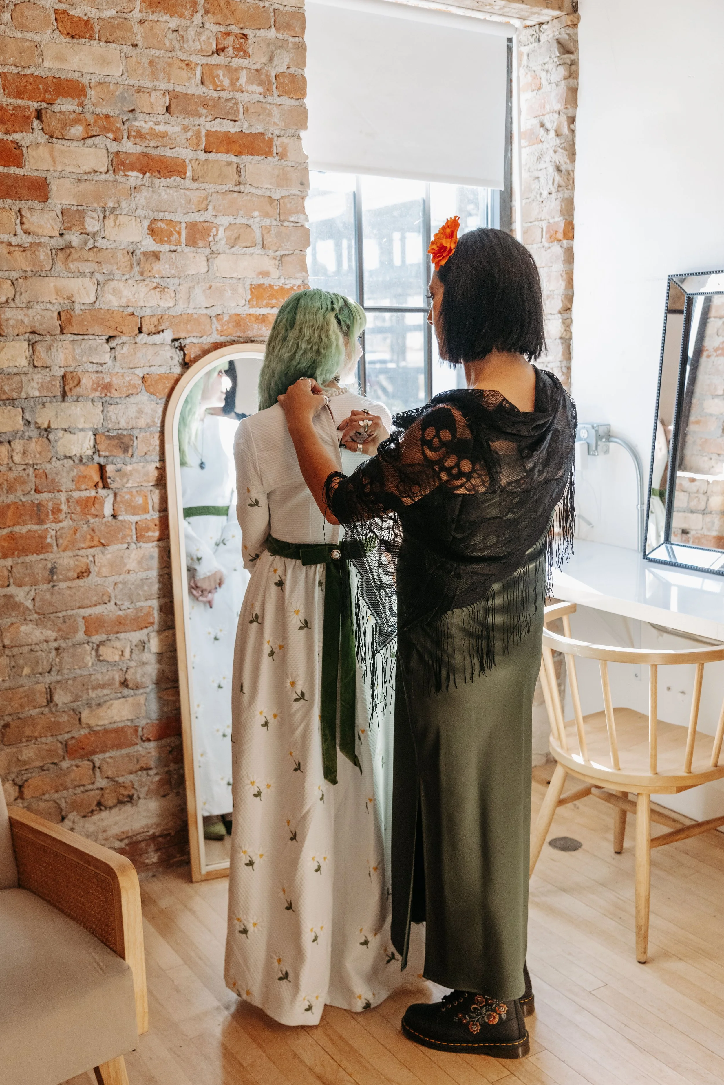 Bride prepares for her wedding while a family member adjusts her dress in a bright bridal suite with exposed brick walls and natural light.