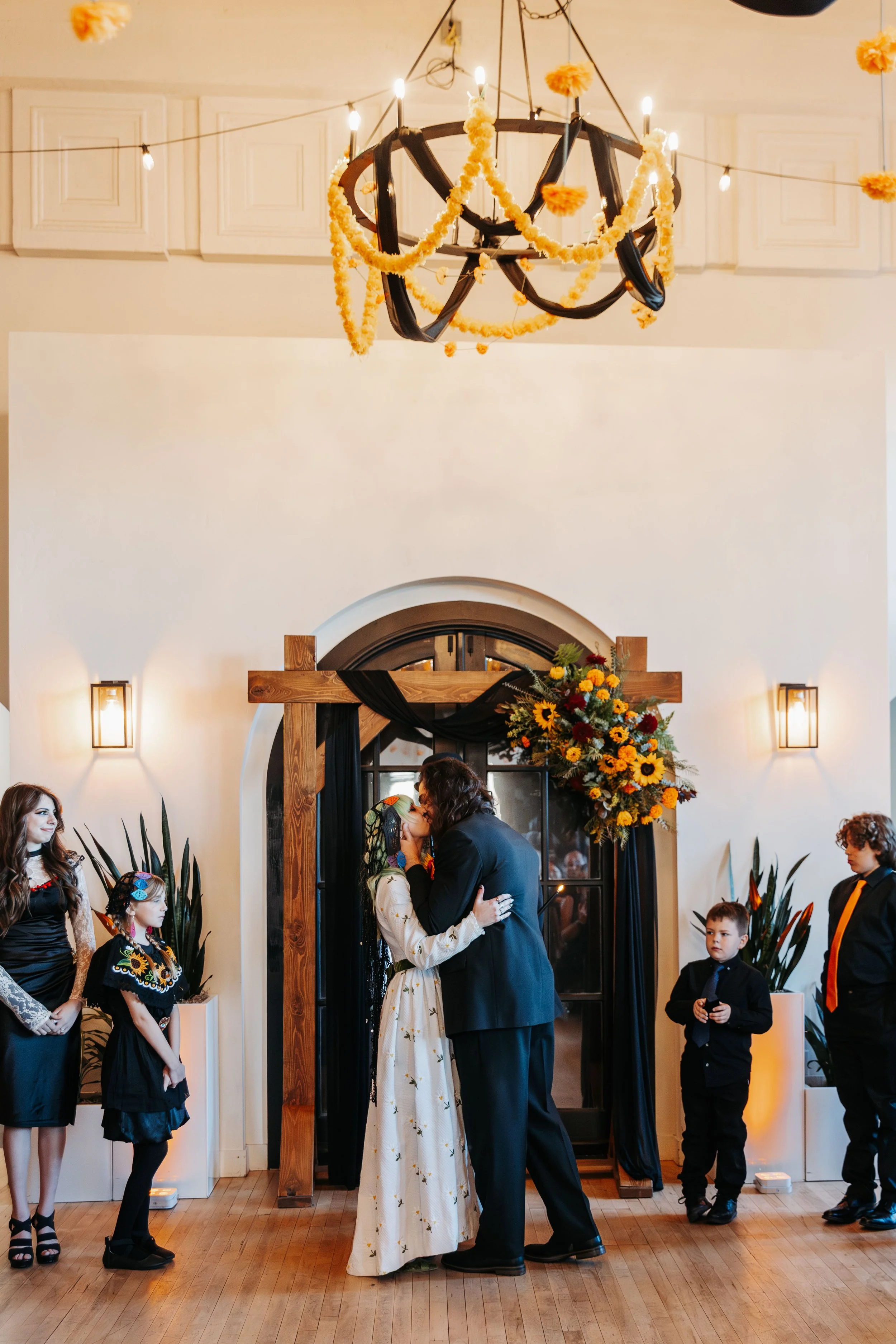 Bride and groom share their first kiss beneath a wooden ceremony arbor decorated with black draping and sunflower-inspired floral arrangements.