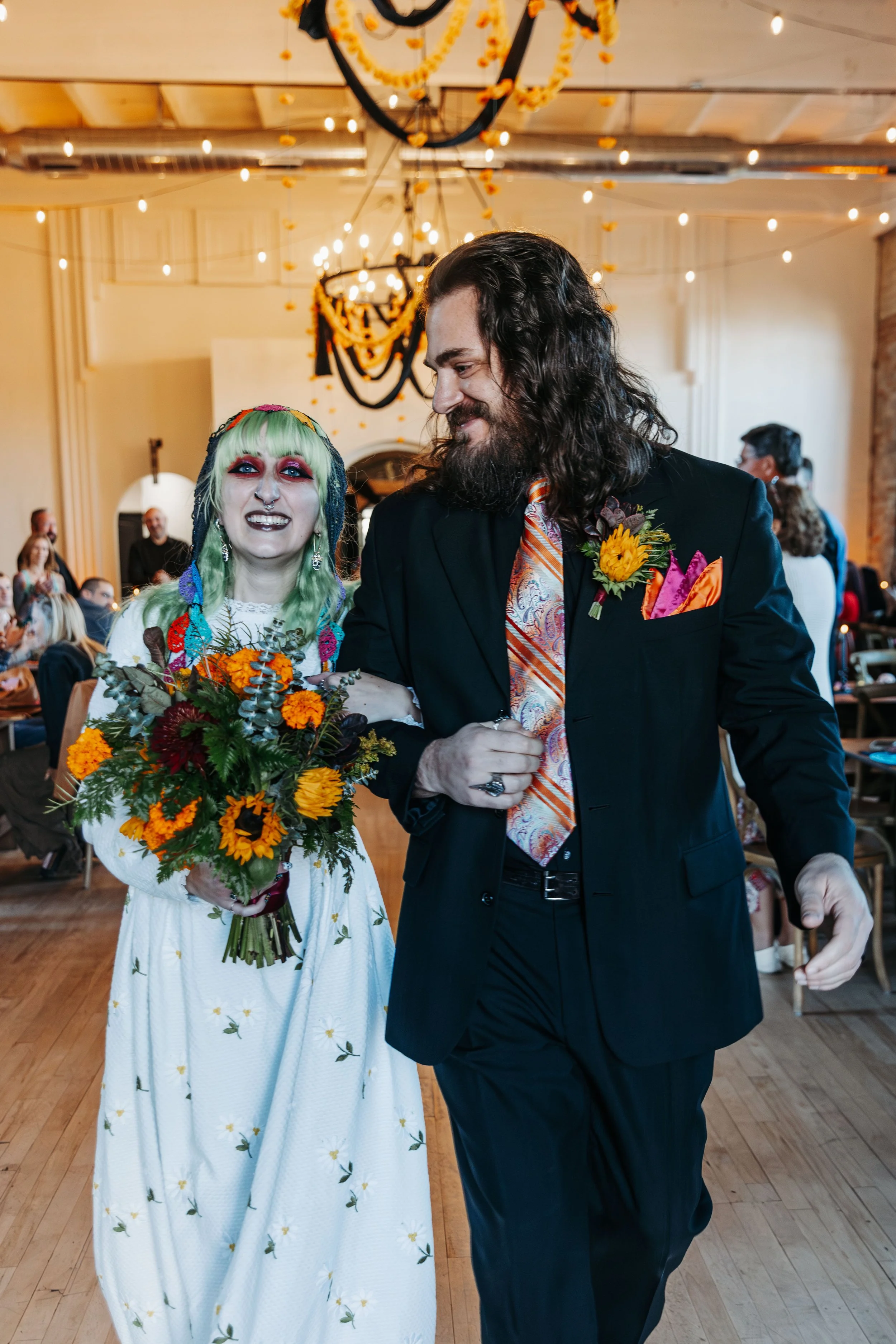 Newly married couple walk arm in arm down the aisle after their ceremony, smiling as guests celebrate around them.