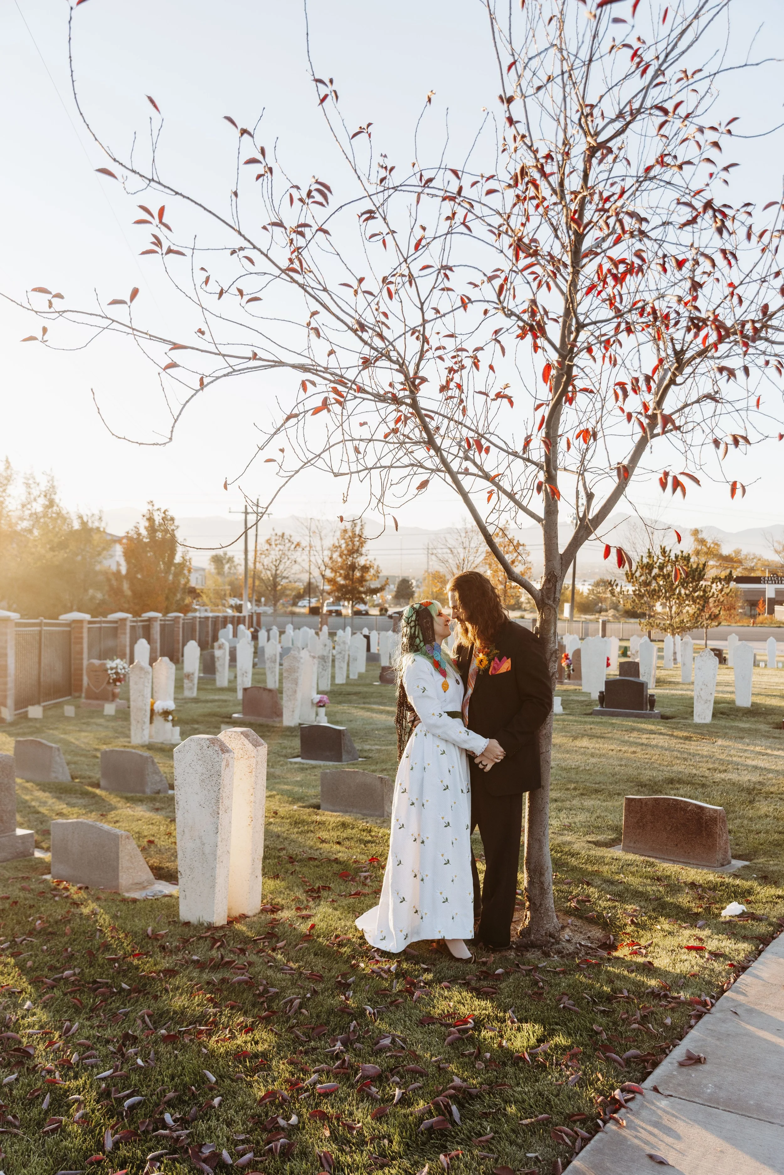 Couple standing beneath a tree in a cemetery during golden hour portraits, reflecting their alternative Día de los Muertos wedding theme in Sandy, Utah.