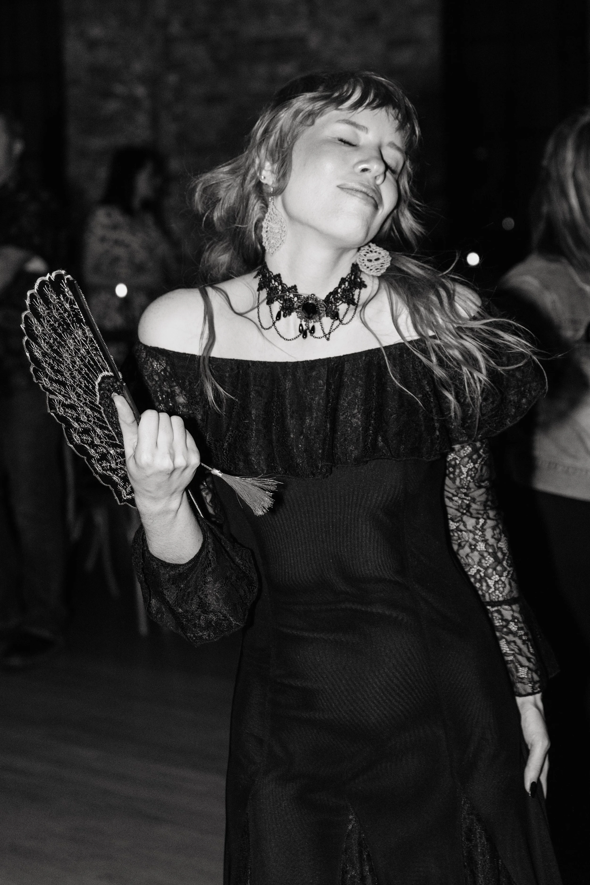 Wedding guest in a black gothic-inspired dress dances with eyes closed while holding a decorative lace fan during the reception.