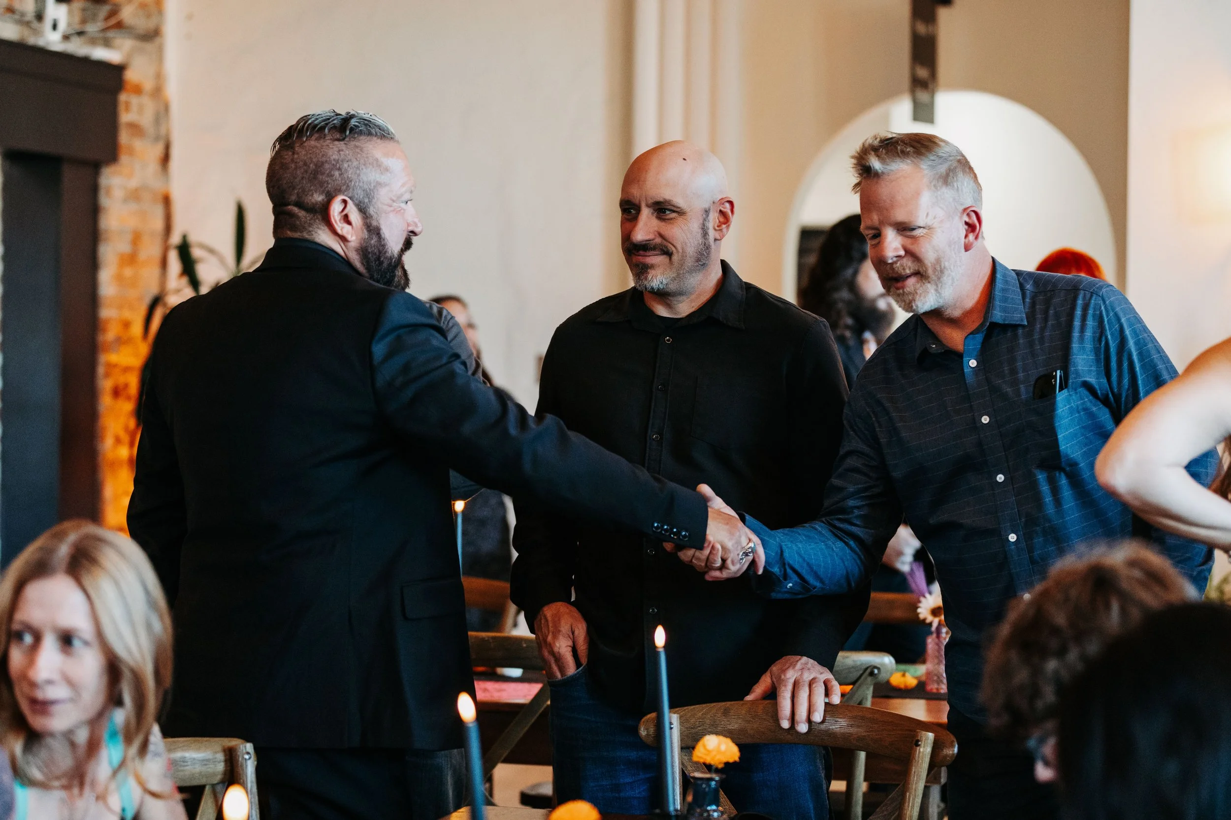 Groom in a black suit shakes hands with wedding guests during the reception at Crescent Hall, surrounded by warm candlelight and reception décor.