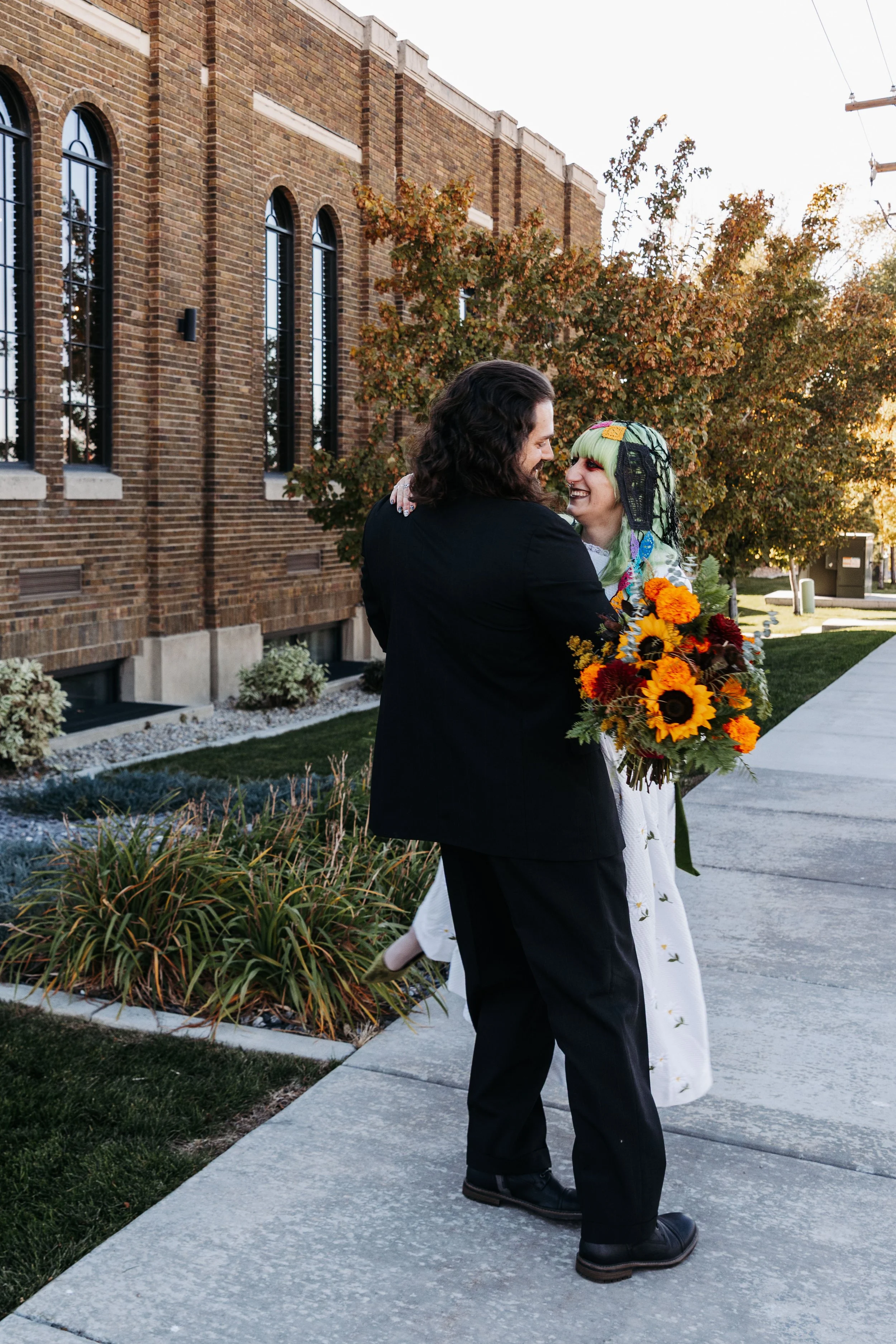 Newlywed couple embraces outside Crescent Hall in Sandy, Utah, with colorful flowers and alternative wedding attire inspired by a Día de los Muertos wedding theme.