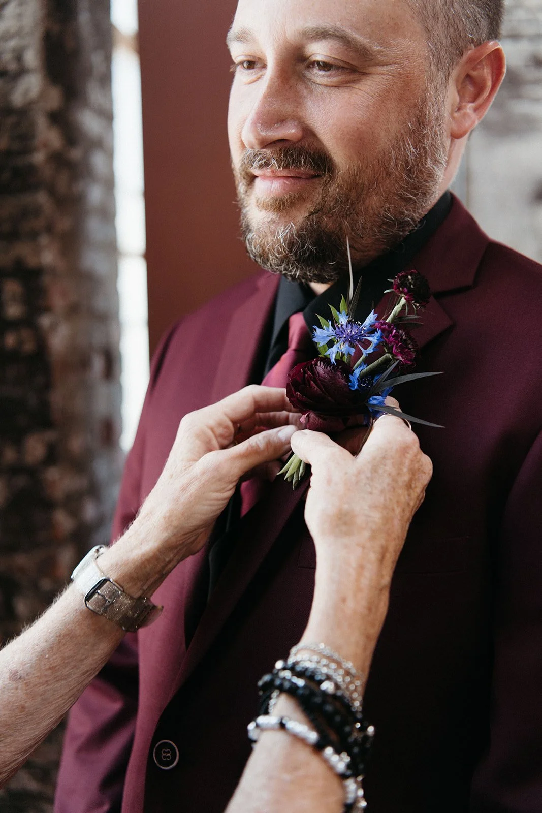 Close-up of a groom in a burgundy suit as a family member pins a colorful floral boutonniere before the wedding ceremony.
