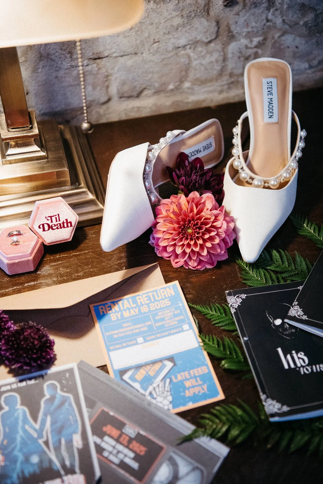 Wedding detail flat lay featuring pearl-trimmed white heels, engagement ring box, themed stationery, florals, and vintage-inspired wedding décor.