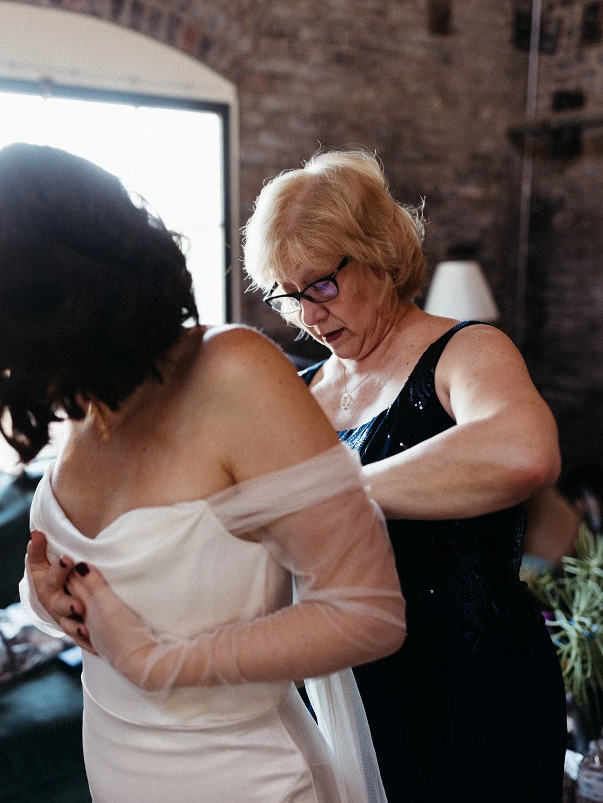 Mother helps adjust the bride's gown during wedding preparations inside a brick loft venue filled with natural light.