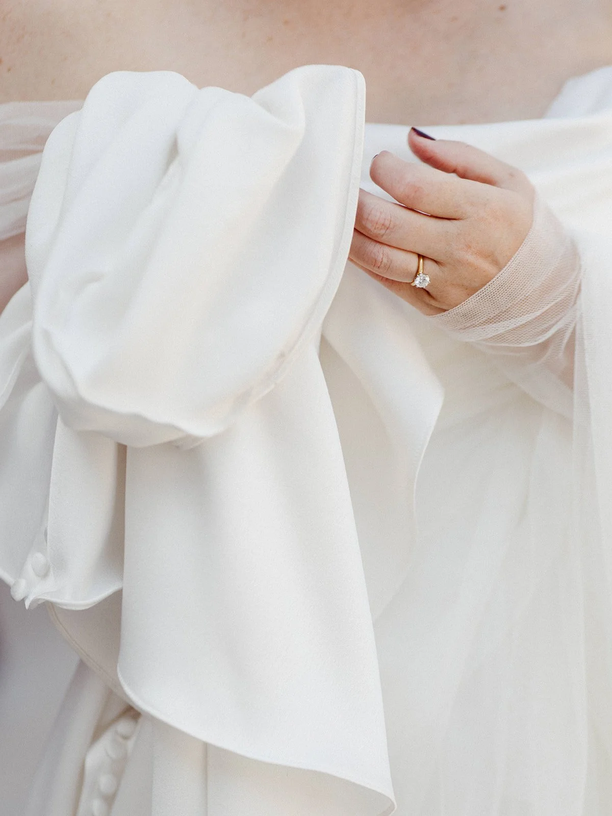 Close-up of the bride's engagement ring and sheer sleeve detail against the draped fabric of her minimalist wedding gown.