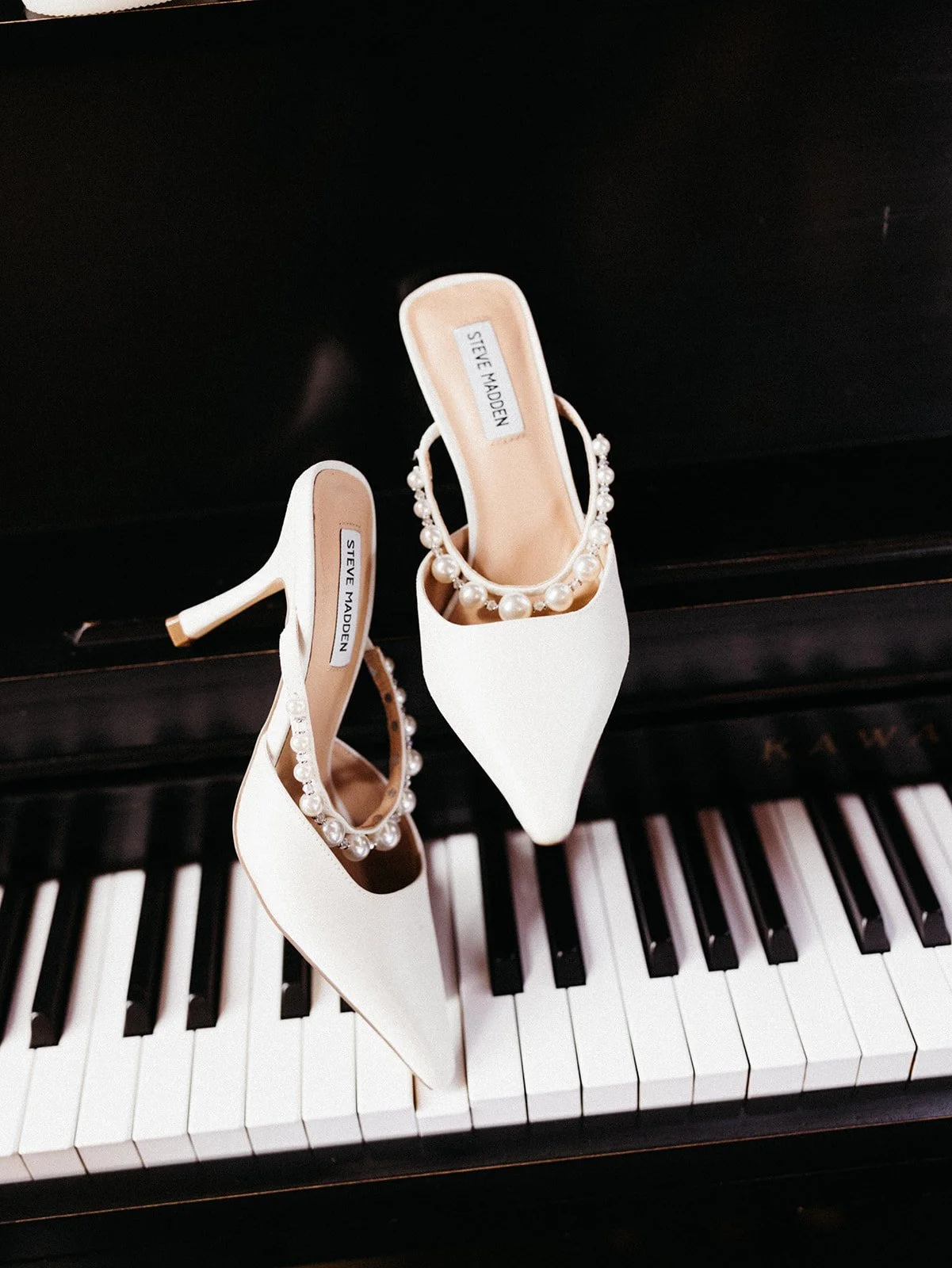 White pearl-embellished bridal heels displayed on the keys of a black piano during wedding detail photography.