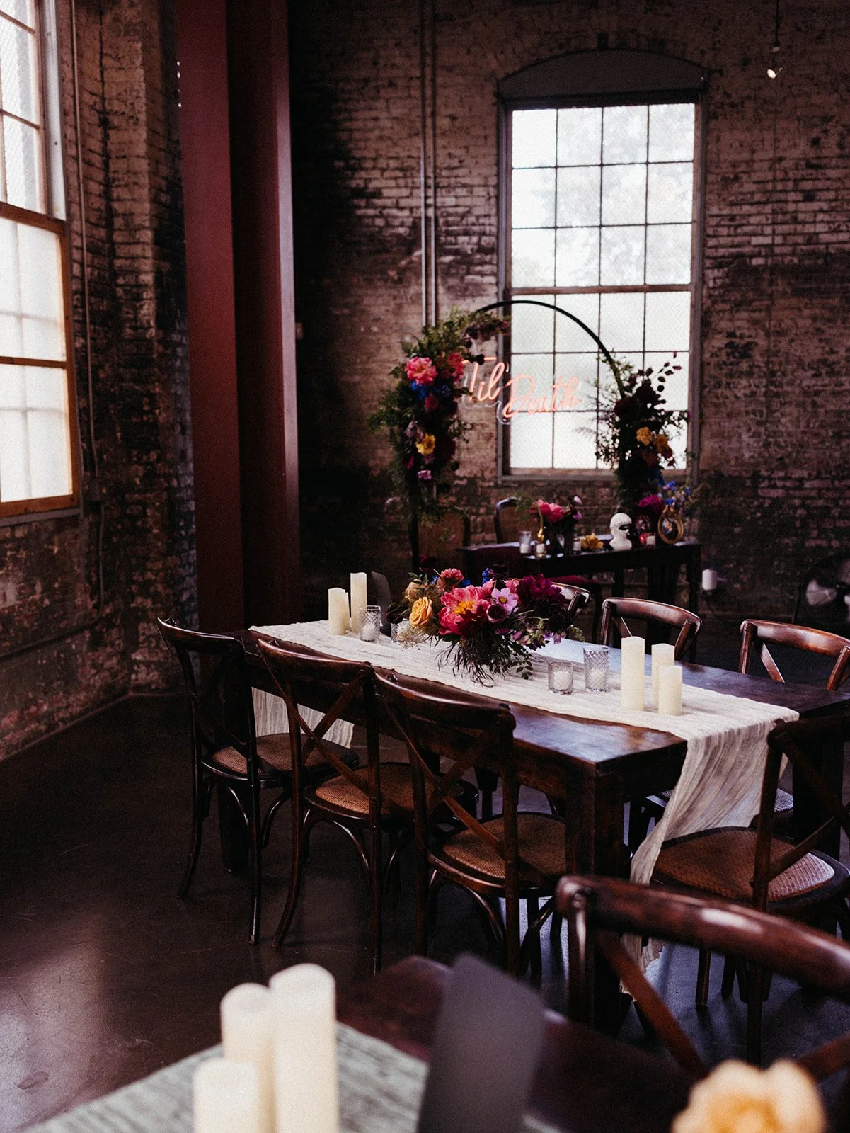Moody wedding reception design featuring floral centerpieces, dark wood tables, and a neon "'Til Death" sign displayed against exposed brick walls.