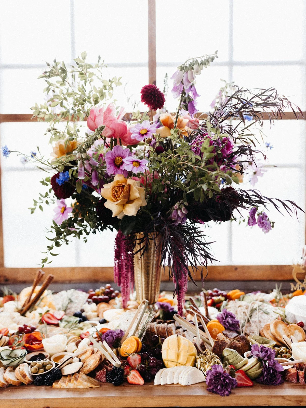 Abundant wedding grazing table featuring artisan cheeses, fruit, charcuterie, and a vibrant floral centerpiece arranged in a gold vase.