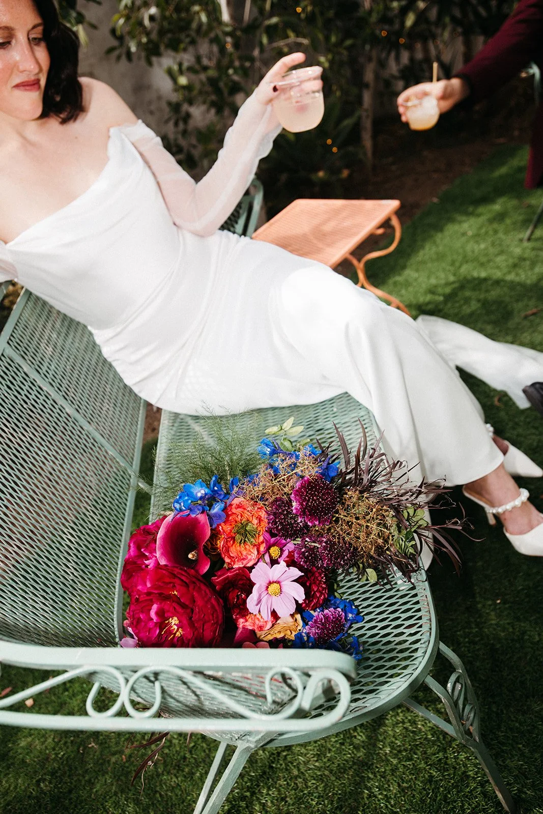 Bride reclining on a vintage metal bench holding a cocktail beside a vibrant bouquet of bold pink, burgundy, and blue wedding flowers.
