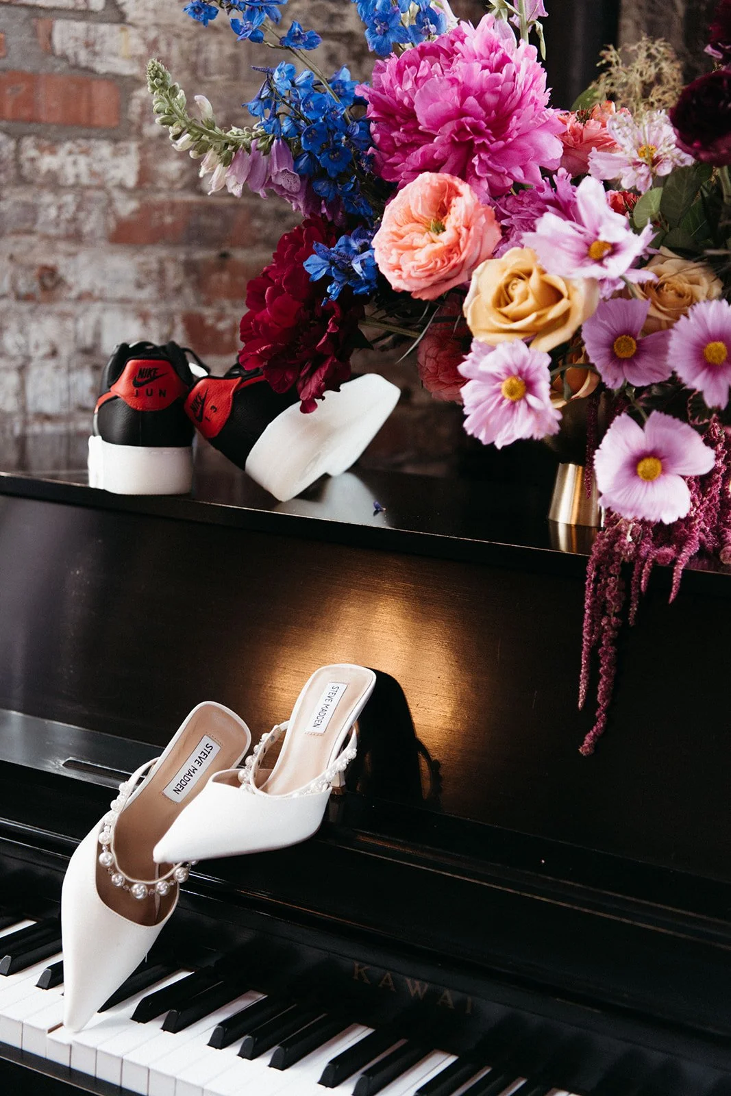Wedding detail styling featuring pearl-trimmed bridal heels, colorful florals, and black-and-red sneakers displayed on a piano.