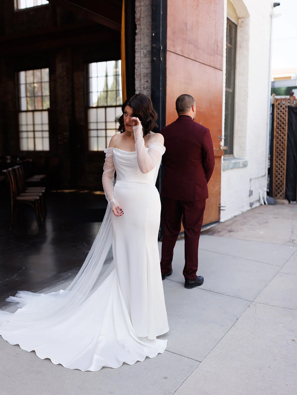 Bride wipes away tears before a first look outside an industrial wedding venue while the groom waits with his back turned.
