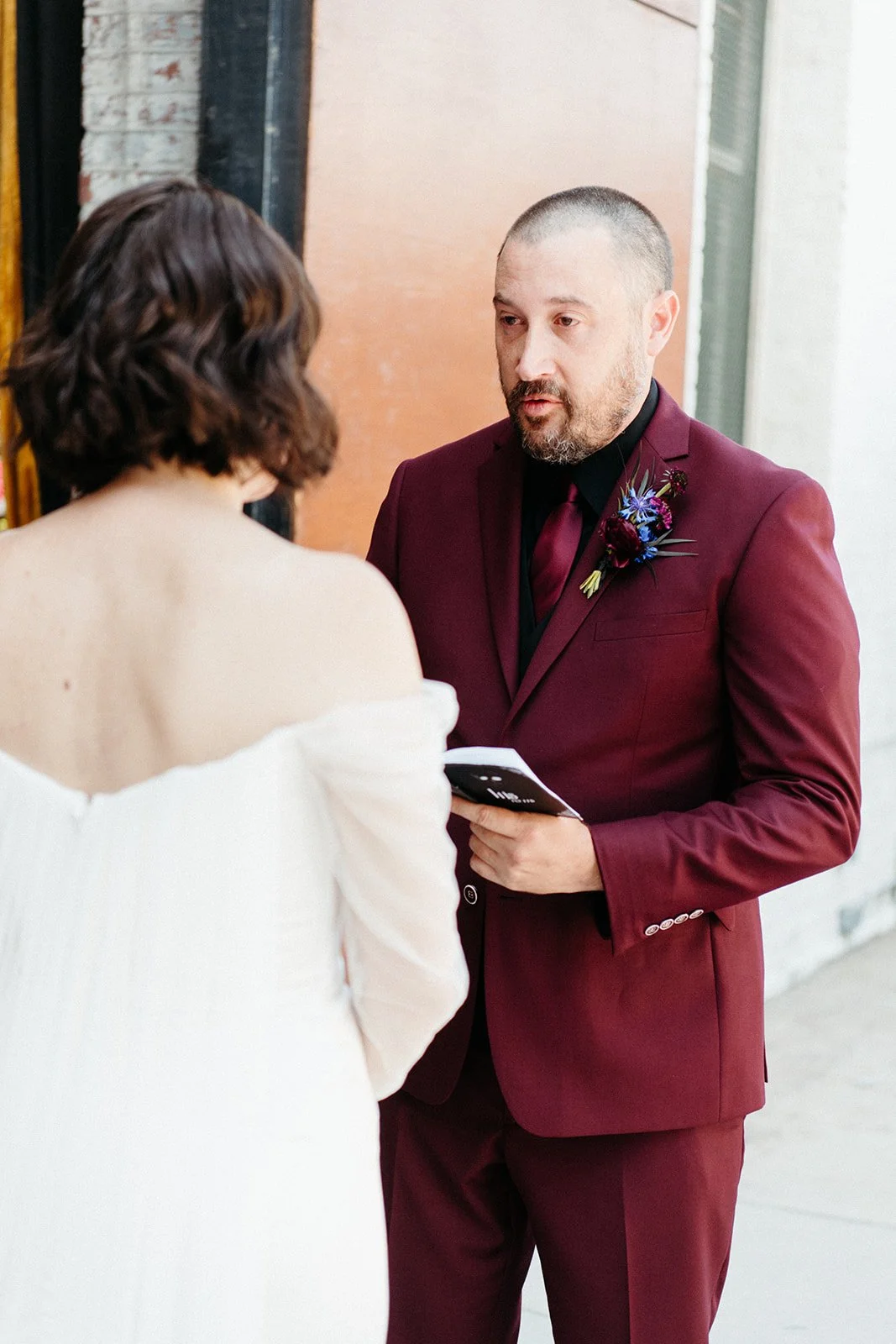 Groom in a burgundy suit reads personal vows to his bride during a private first look outside a historic industrial wedding venue.