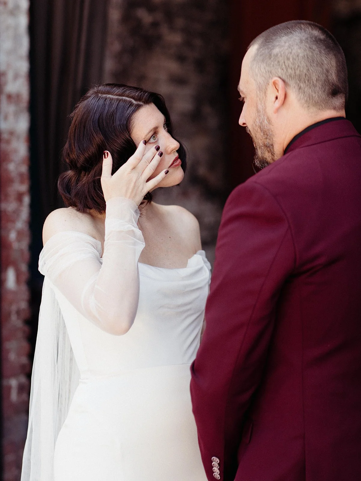 Bride wipes away tears while standing face-to-face with her groom during an emotional first look outside an industrial wedding venue.