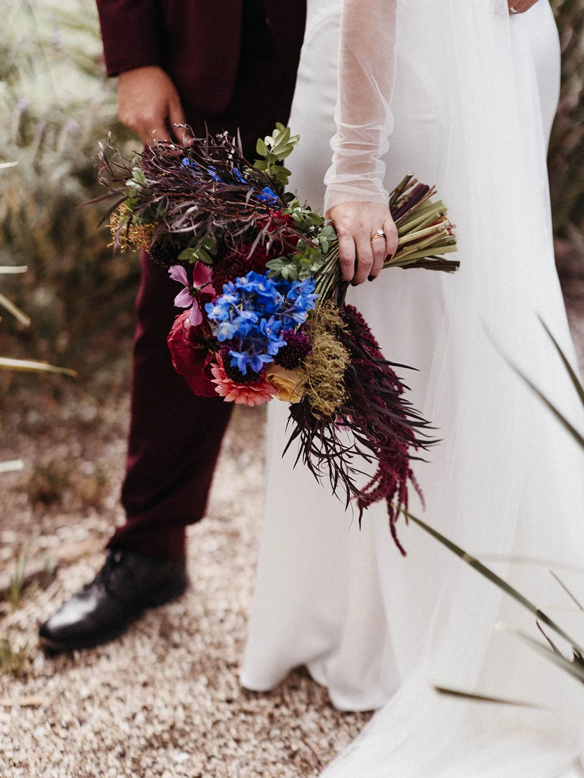 Close-up of a bride holding a dramatic bouquet featuring blue delphinium, burgundy blooms, pink flowers, and textured foliage.