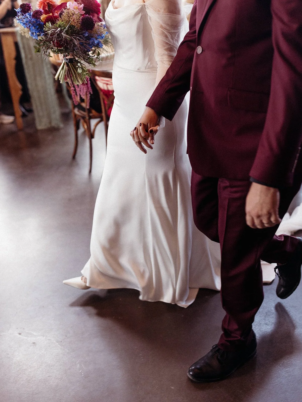 Bride in an off-the-shoulder wedding gown with sheer sleeves admires her reflection in a full-length mirror inside an industrial loft wedding venue.