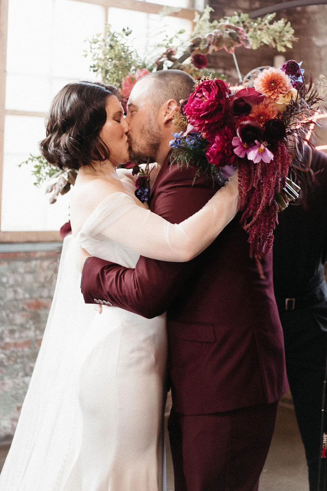 Groom in a burgundy suit kisses the bride as wedding guests applaud during a ceremony at an industrial loft wedding venue.