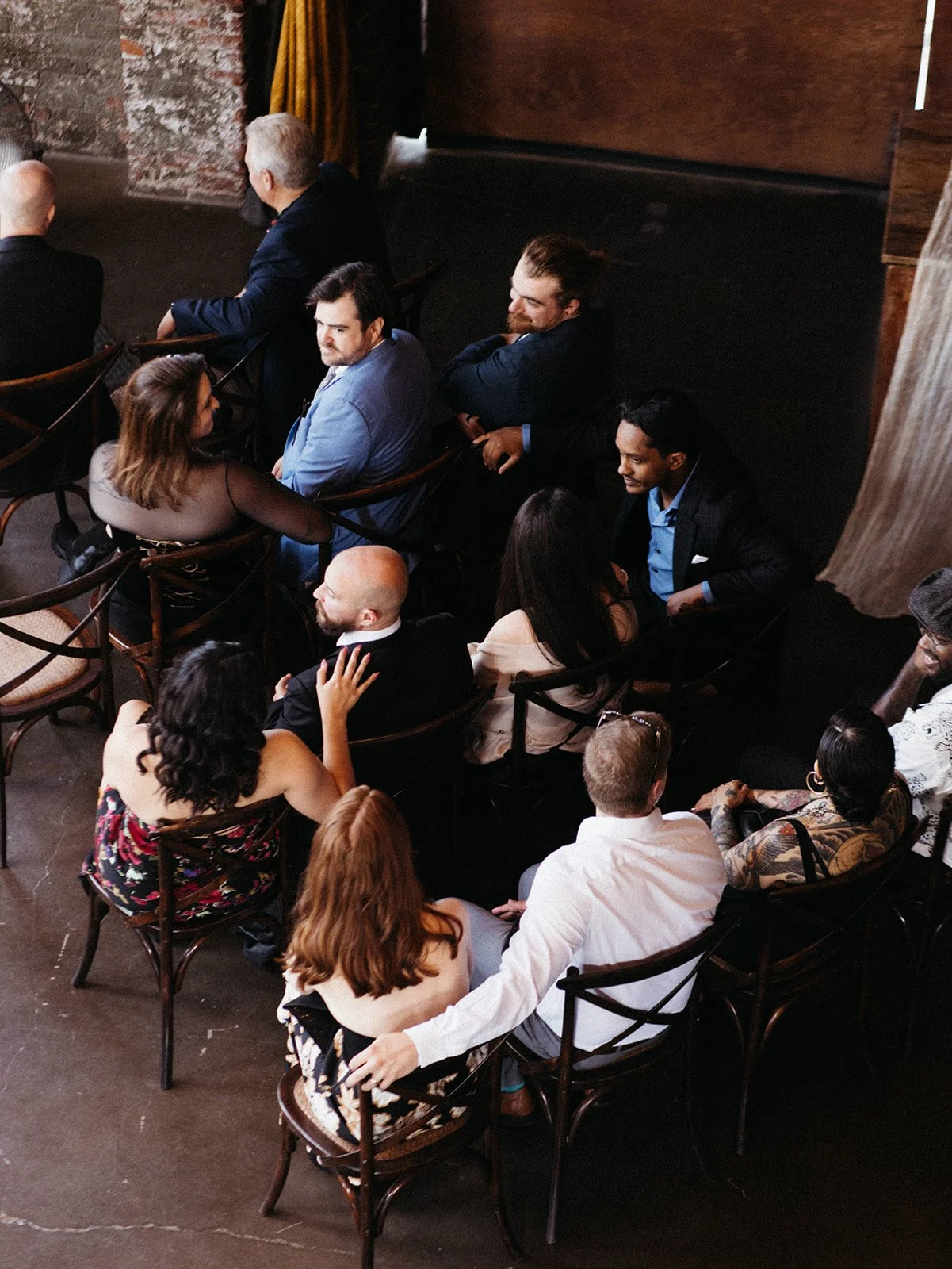 Wedding guests seated in wooden cross-back chairs watch the ceremony inside an industrial loft venue with exposed brick walls and warm natural light.