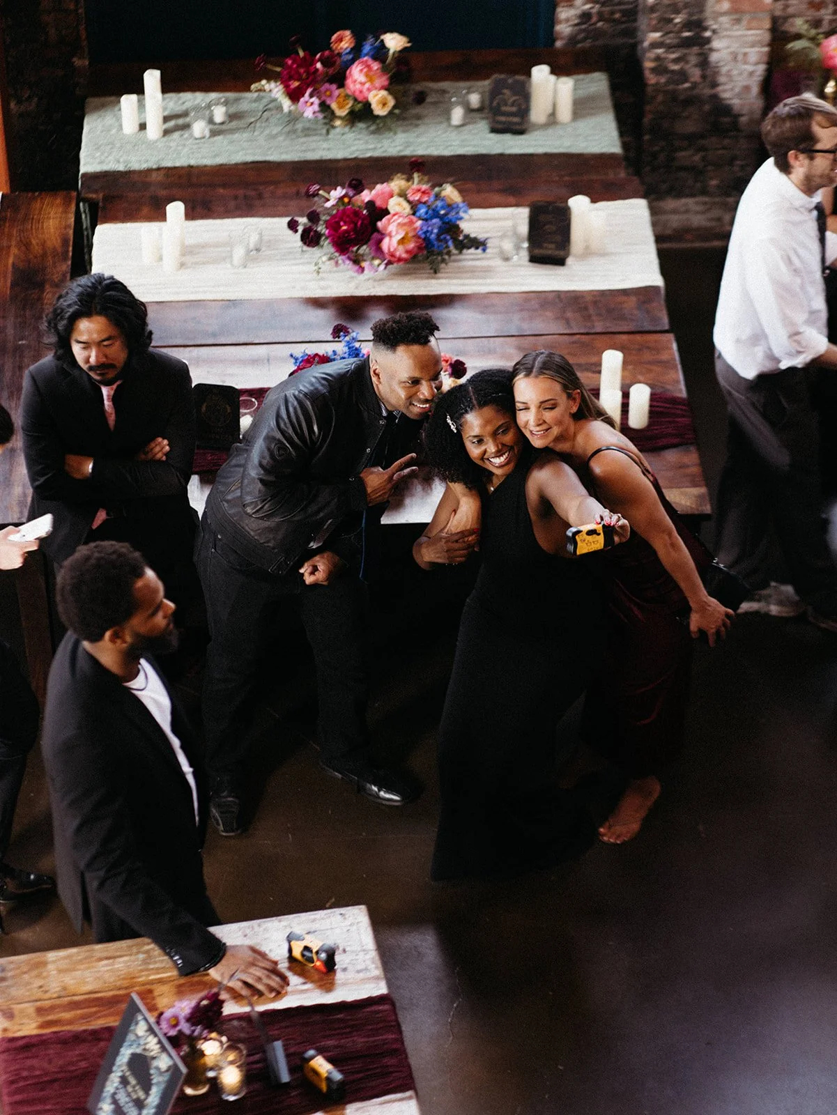 Wedding guests gather around a reception table to take a candid selfie, surrounded by colorful floral centerpieces and rustic wooden tables.